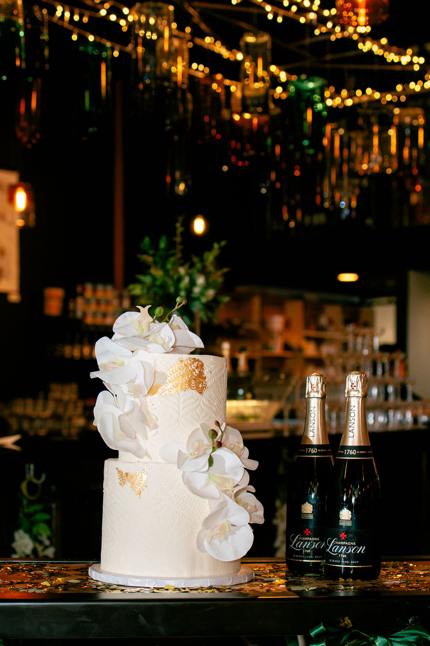 Elegant two-tier white wedding cake with gold leaf and orchids beside two champagne bottles in a dark venue.