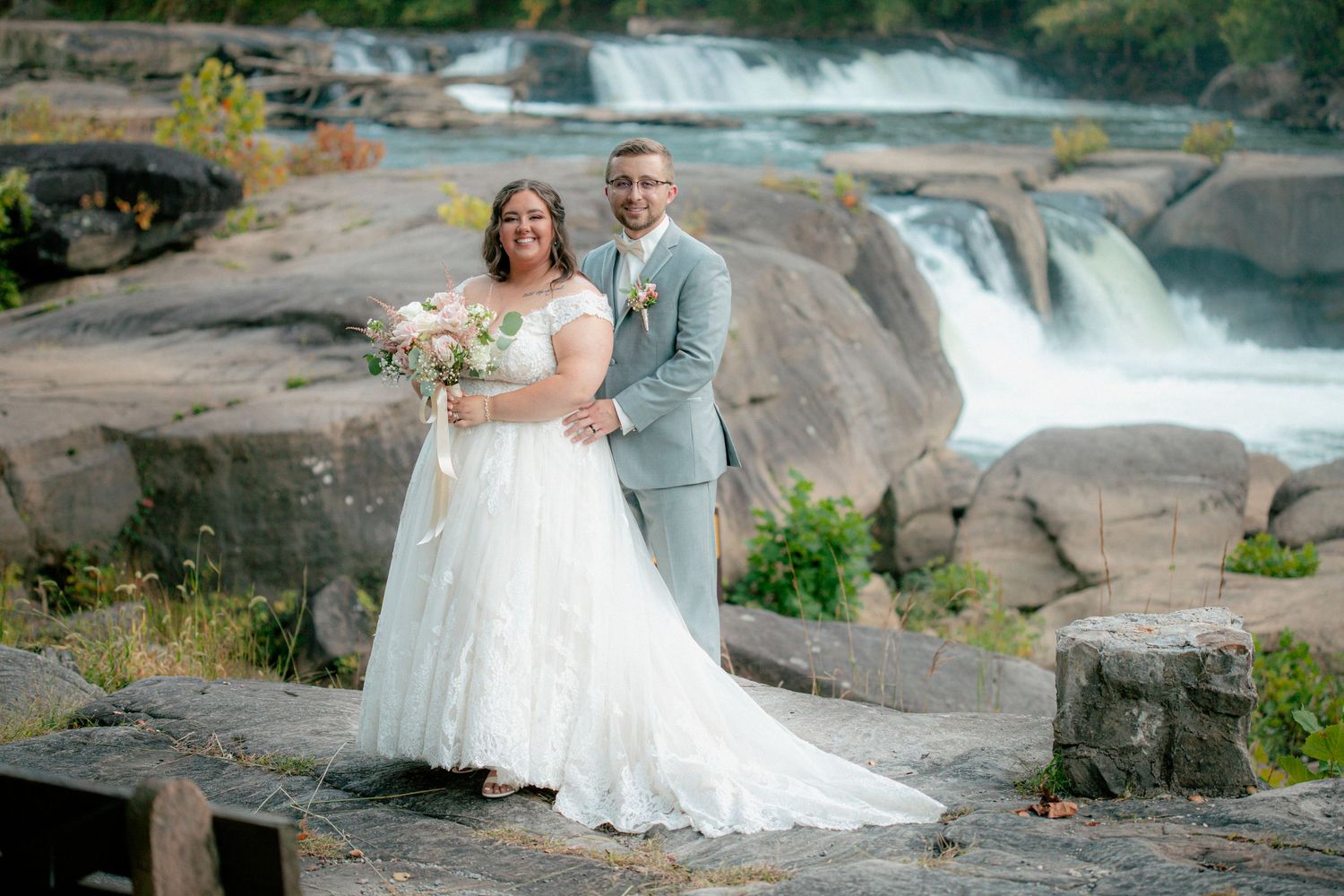 Bride and groom smile together holding pink bouquet with dramatic waterfall cascading over rocky cliffs in the background.