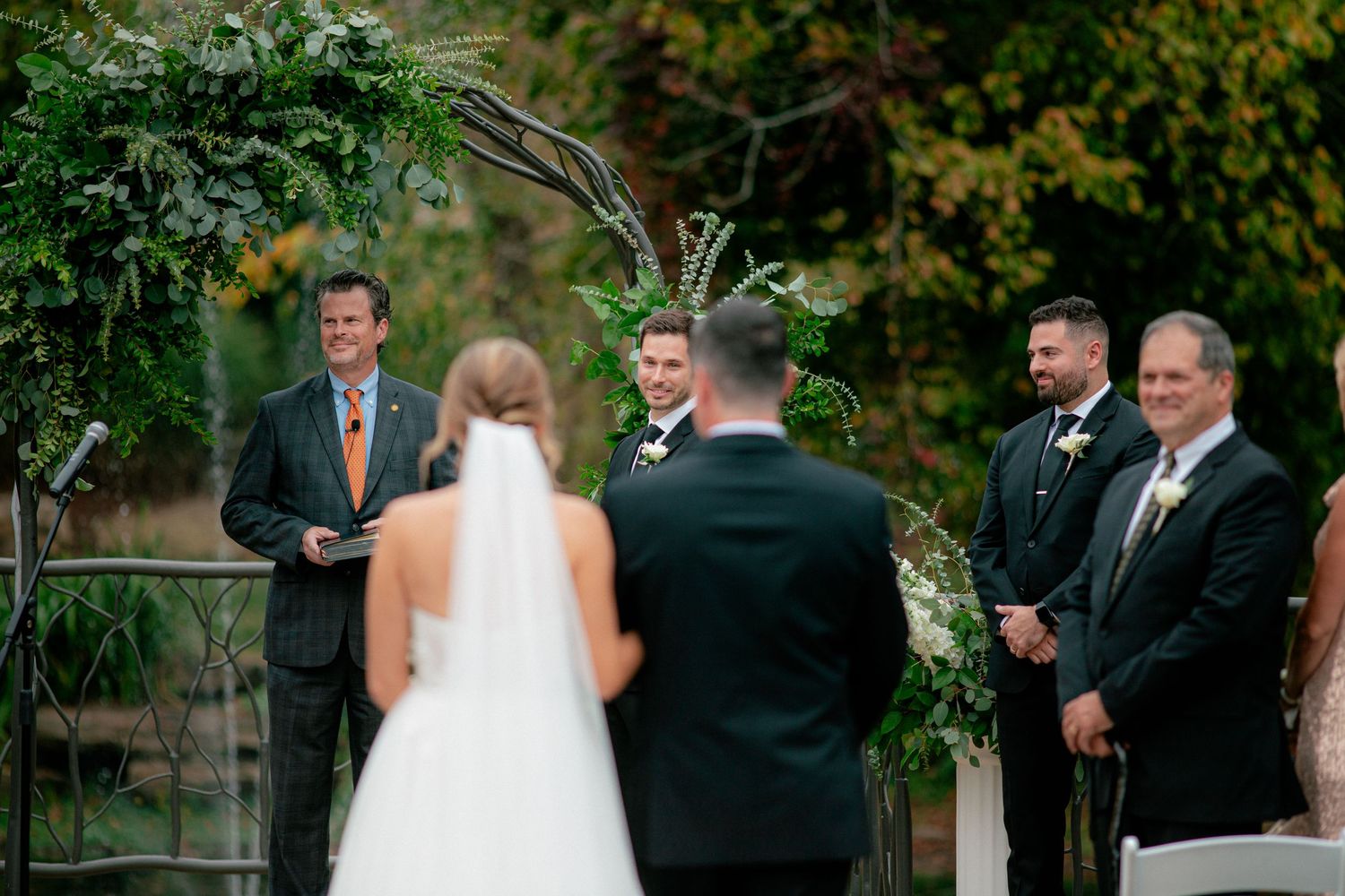 Groom smiling as bride walks down the aisle at an outdoor wedding ceremony with lush greenery arch.