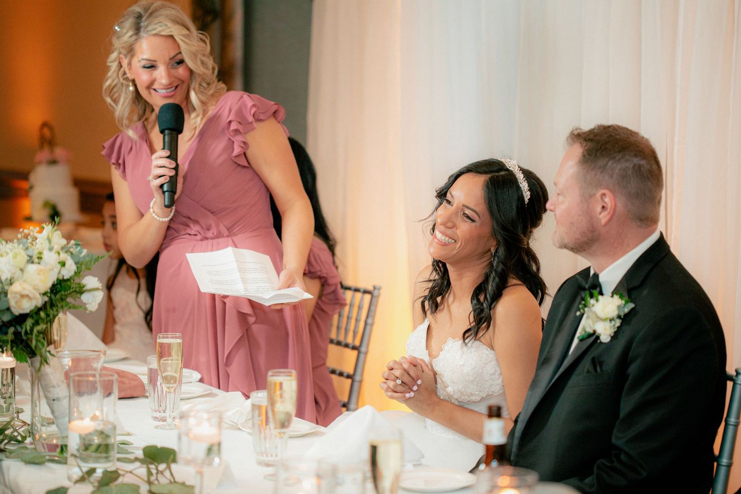 Bridesmaid in pink dress giving heartfelt speech to smiling bride and groom at reception.