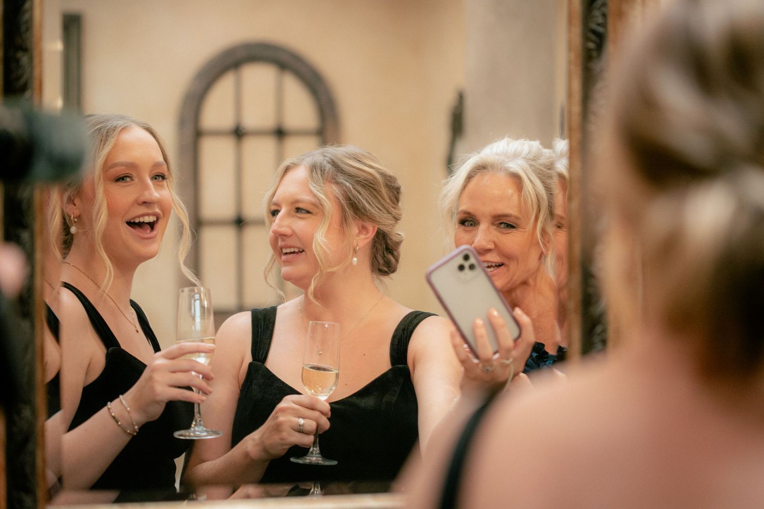 Bridesmaids in black dresses laughing and taking a selfie while holding champagne glasses in front of a mirror.