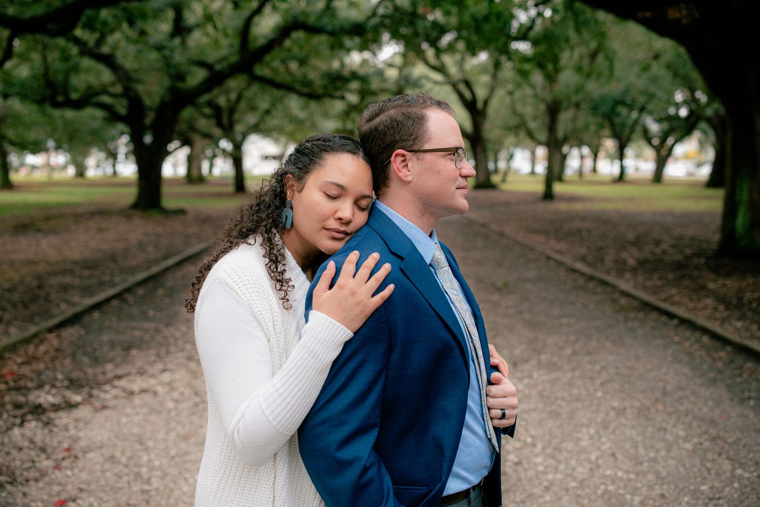 Woman in white sweater rests her head on mans back in blue suit along a tree-lined path.