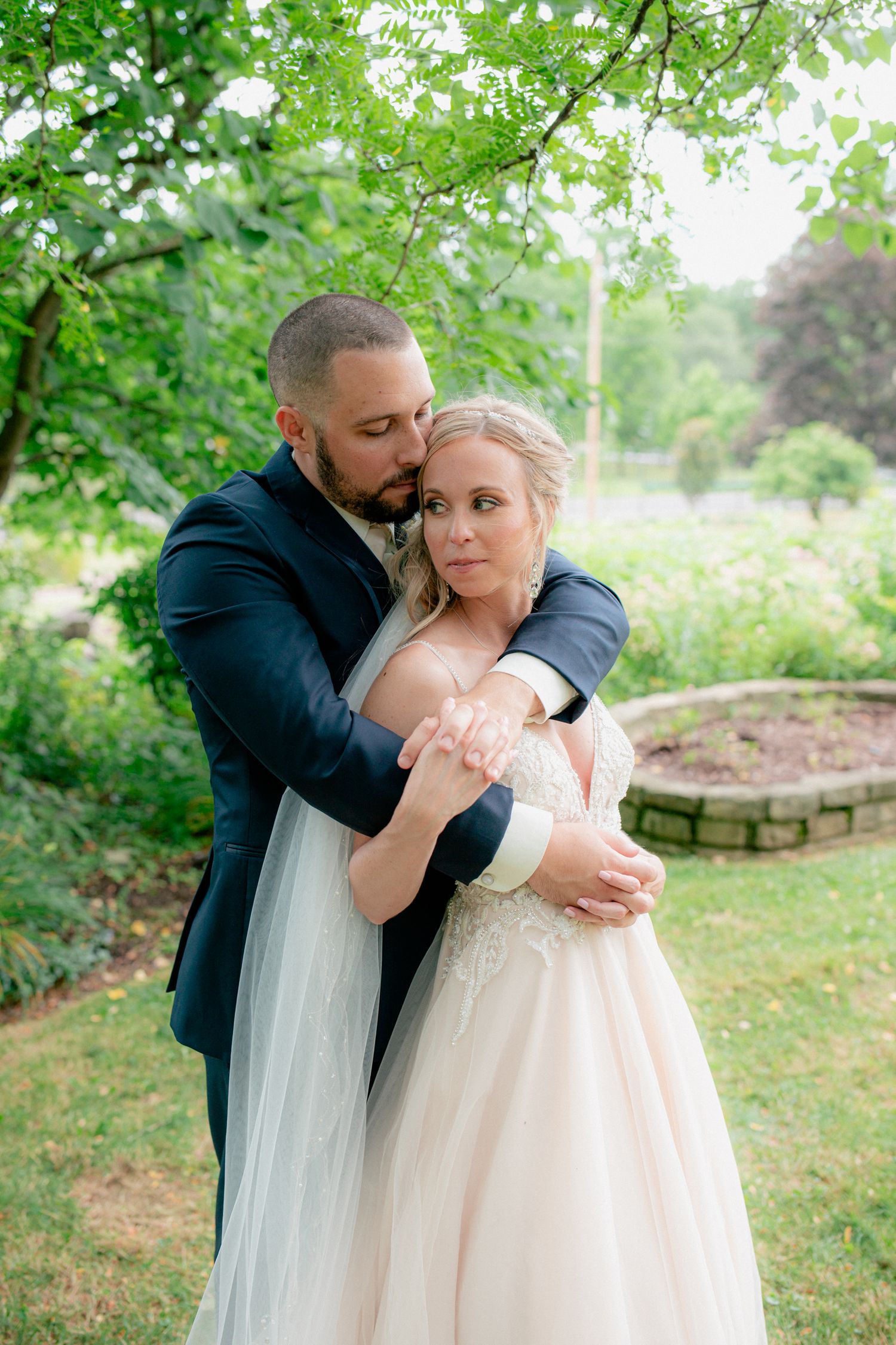 Groom in navy suit embracing bride in blush gown from behind in a garden setting.
