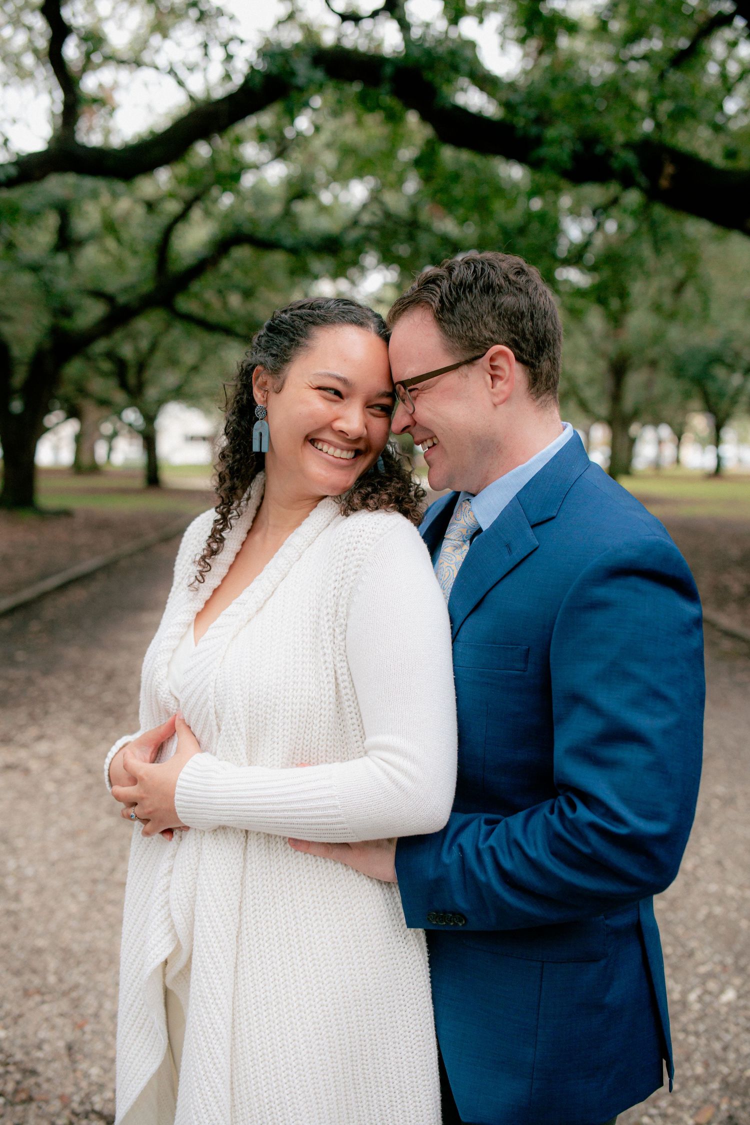 Joyful couple laughs together outdoors under oak trees, bride in white knit dress and groom in blue suit.