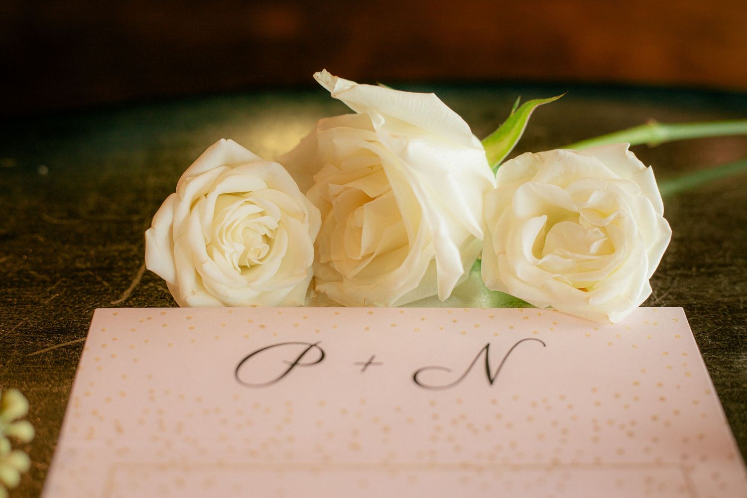 Three white roses resting on a pink card inscribed with P plus N for wedding stationery.