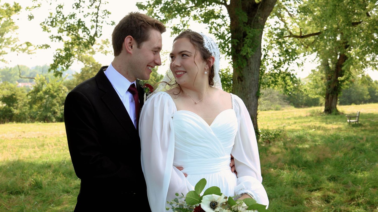 Newlywed couple sharing a joyful glance outdoors surrounded by green trees, bride in white gown with veil.