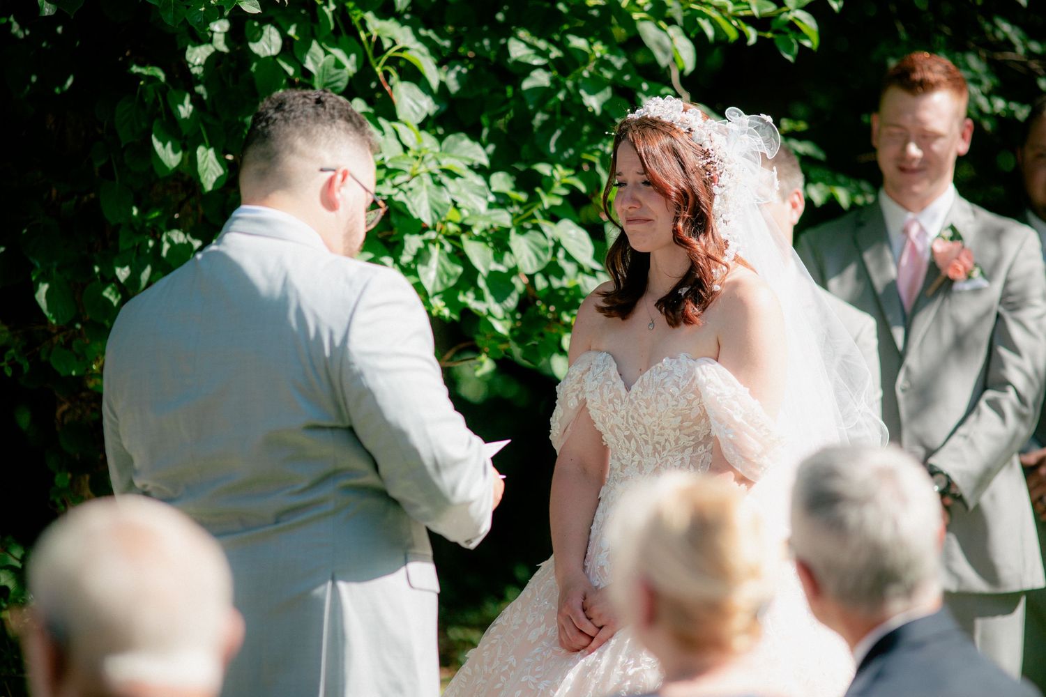 Bride and groom exchange vows outdoors under green foliage with a smiling officiant nearby.