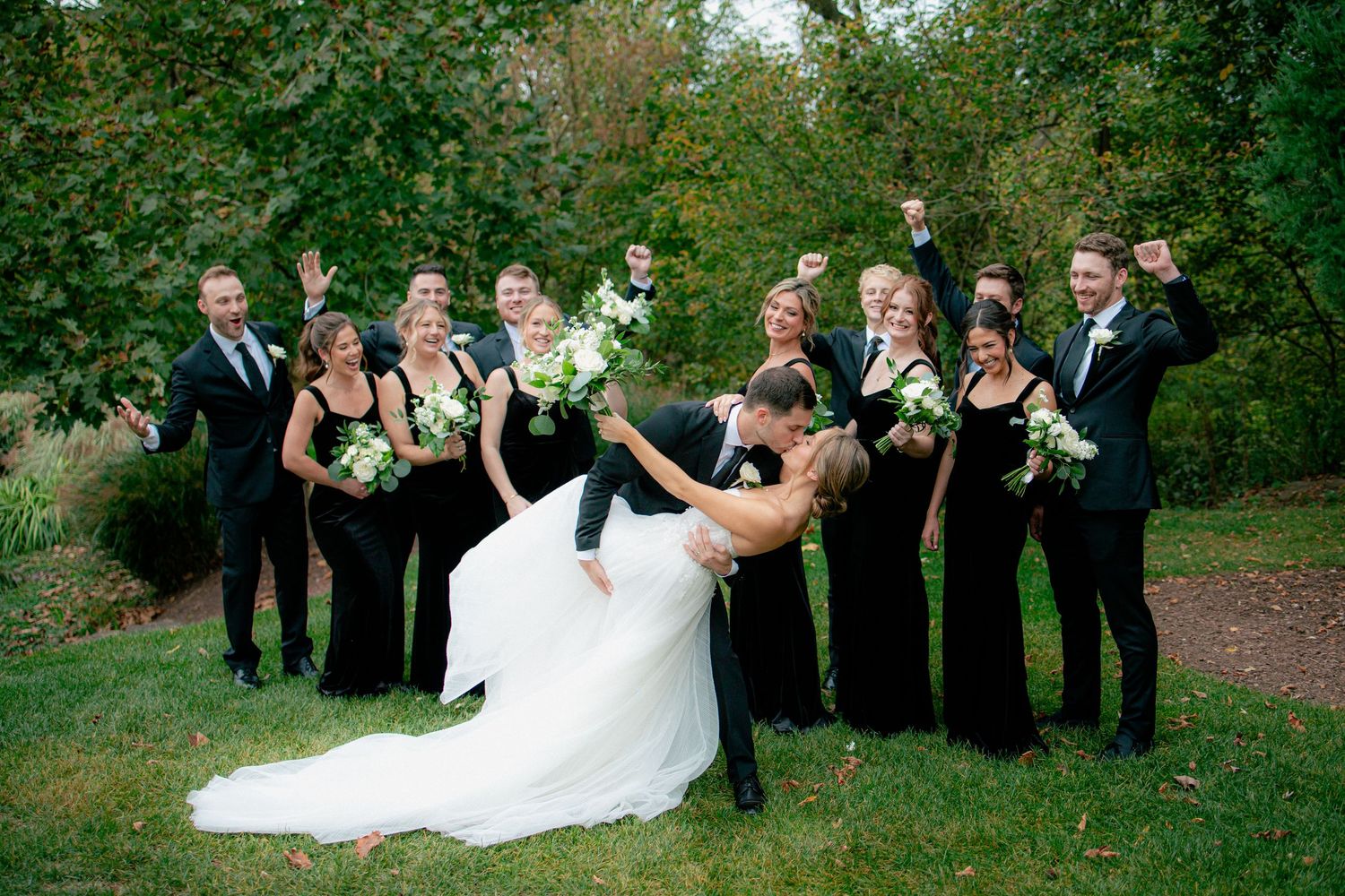 Wedding party celebrates as groom dips bride for a kiss, bridesmaids in black gowns and groomsmen in suits cheering outdoors.