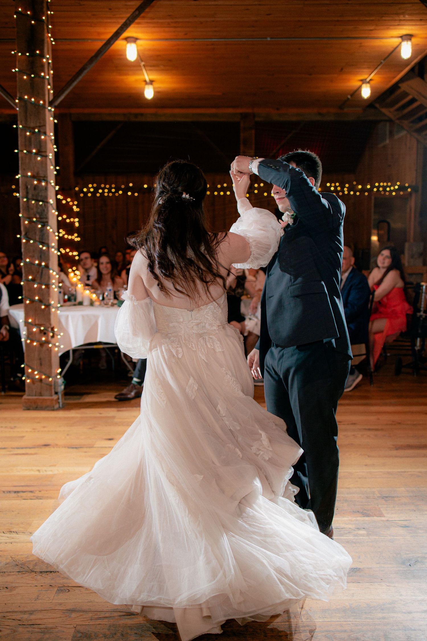 Bride and groom twirl together during their first dance in a rustic barn venue with fairy lights.
