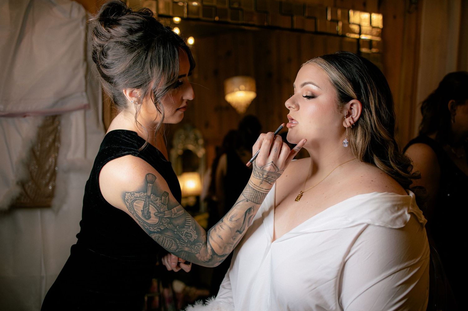 Tattooed makeup artist applying lipstick to a bride in an off-shoulder white wedding gown.