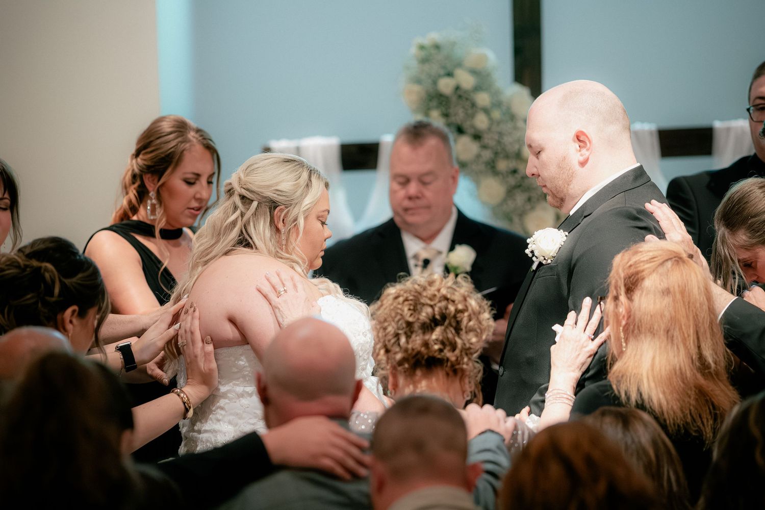 Wedding guests gather around bride and groom for group prayer during heartfelt ceremony moment.