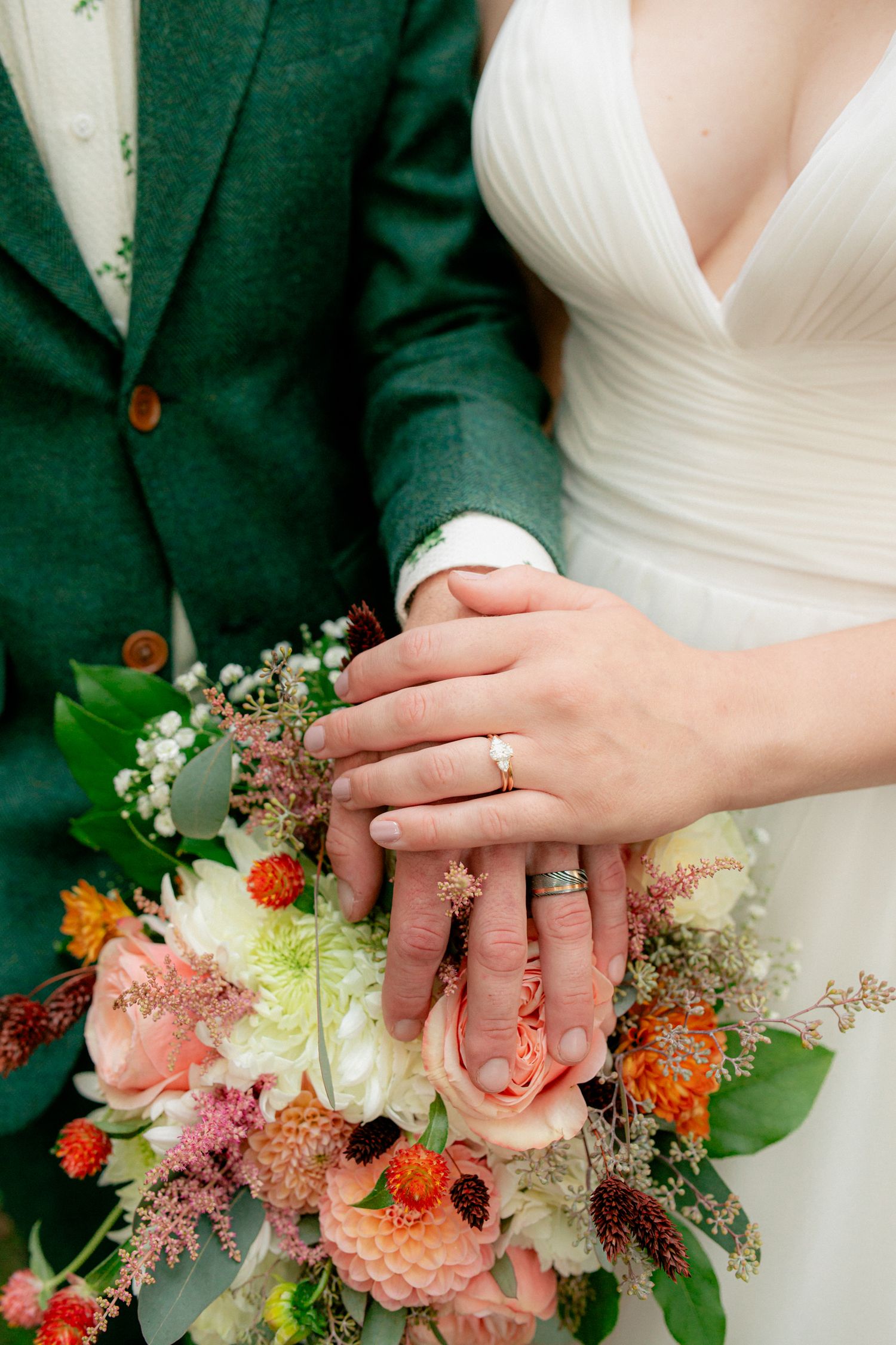 Bride and groom displaying wedding rings on a colorful bridal bouquet with peach and orange flowers.