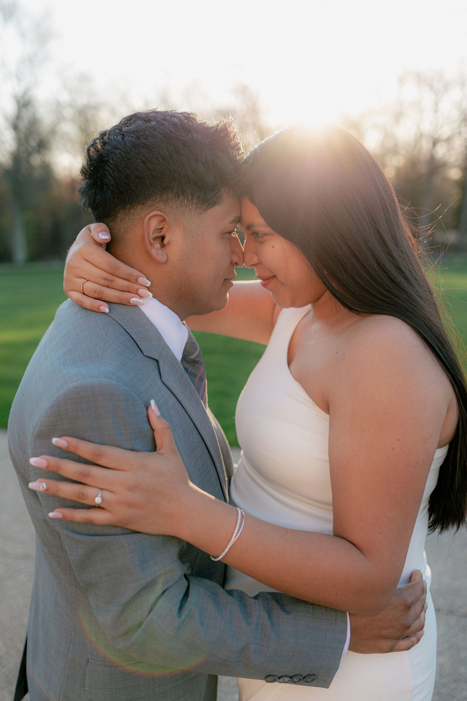 Couple embracing forehead-to-forehead in golden sunset light, bride in white dress and groom in gray suit.