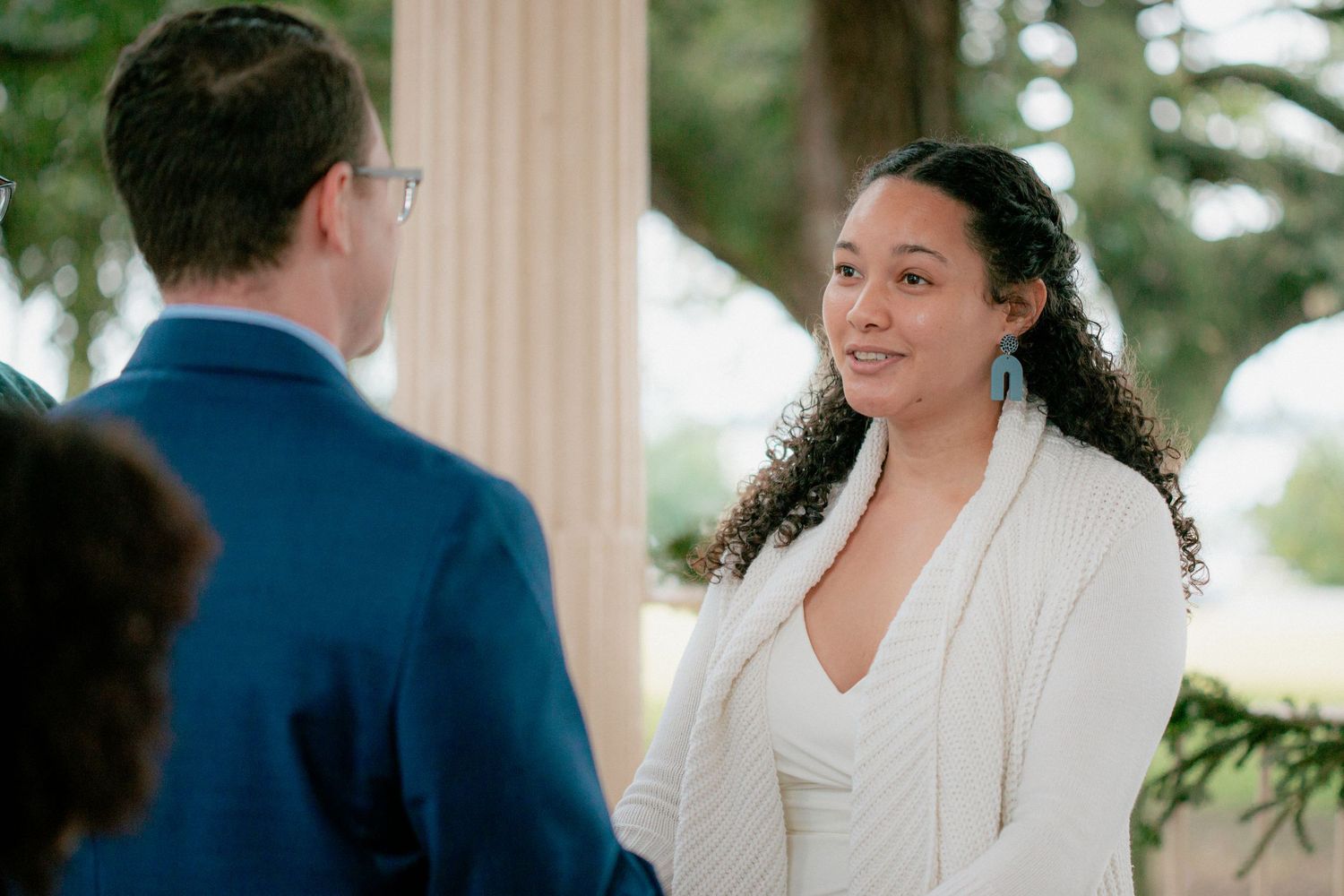 Radiant bride in white knit wrap smiles at groom during outdoor wedding ceremony under oak trees.