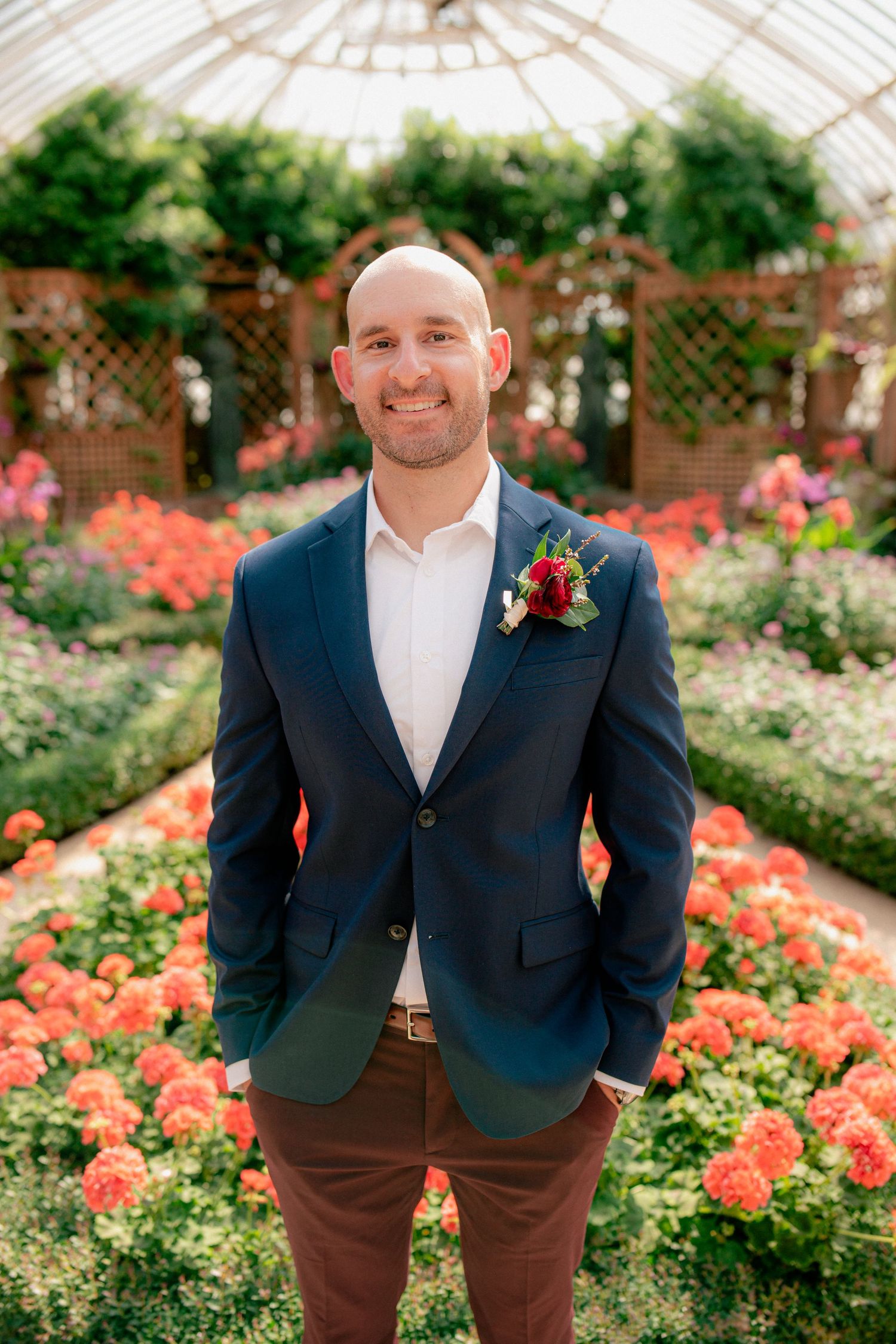 Smiling bald groom in navy blazer with boutonniere stands in a flower greenhouse.