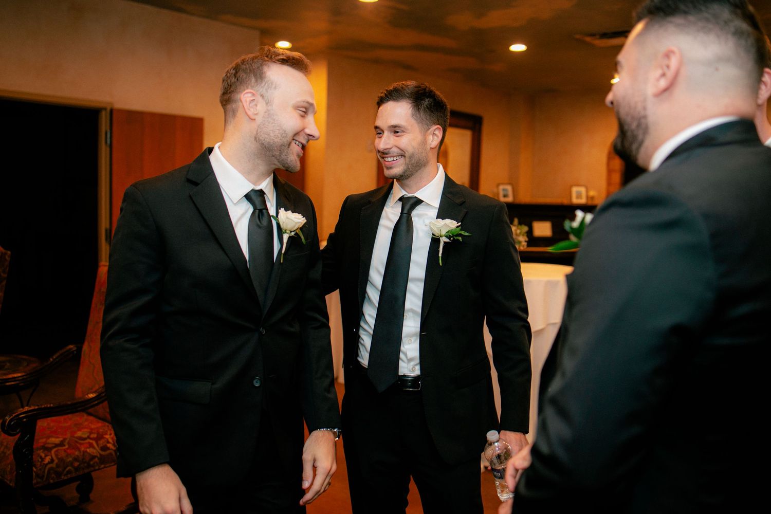 Groomsmen in black suits smiling and conversing before a wedding ceremony indoors.