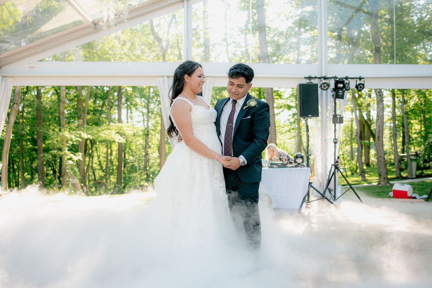 Bride and groom share first dance on cloud-filled floor under elegant tent surrounded by trees.
