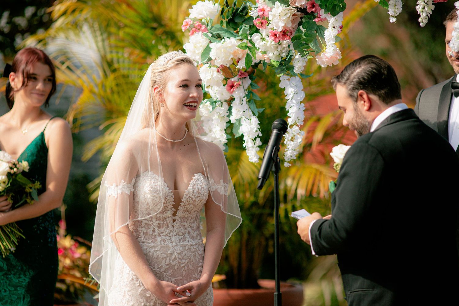 Smiling blonde bride in lace gown exchanges vows under floral arch at tropical wedding ceremony.