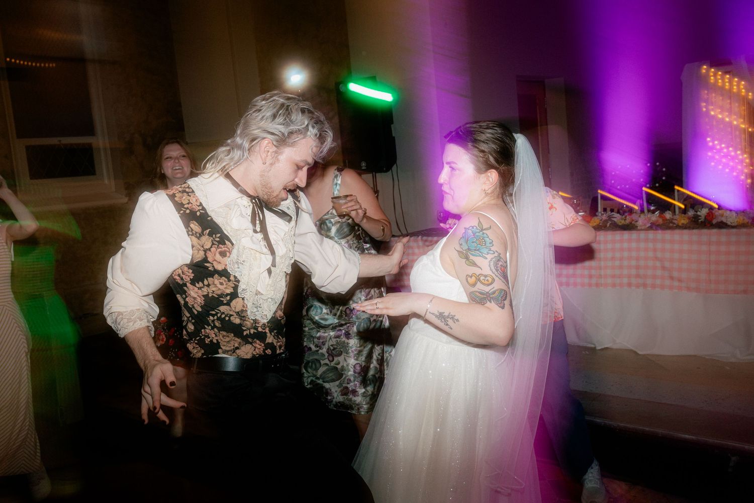 Bride in white gown and veil dancing with a guest in floral vest at a wedding reception with purple lighting.
