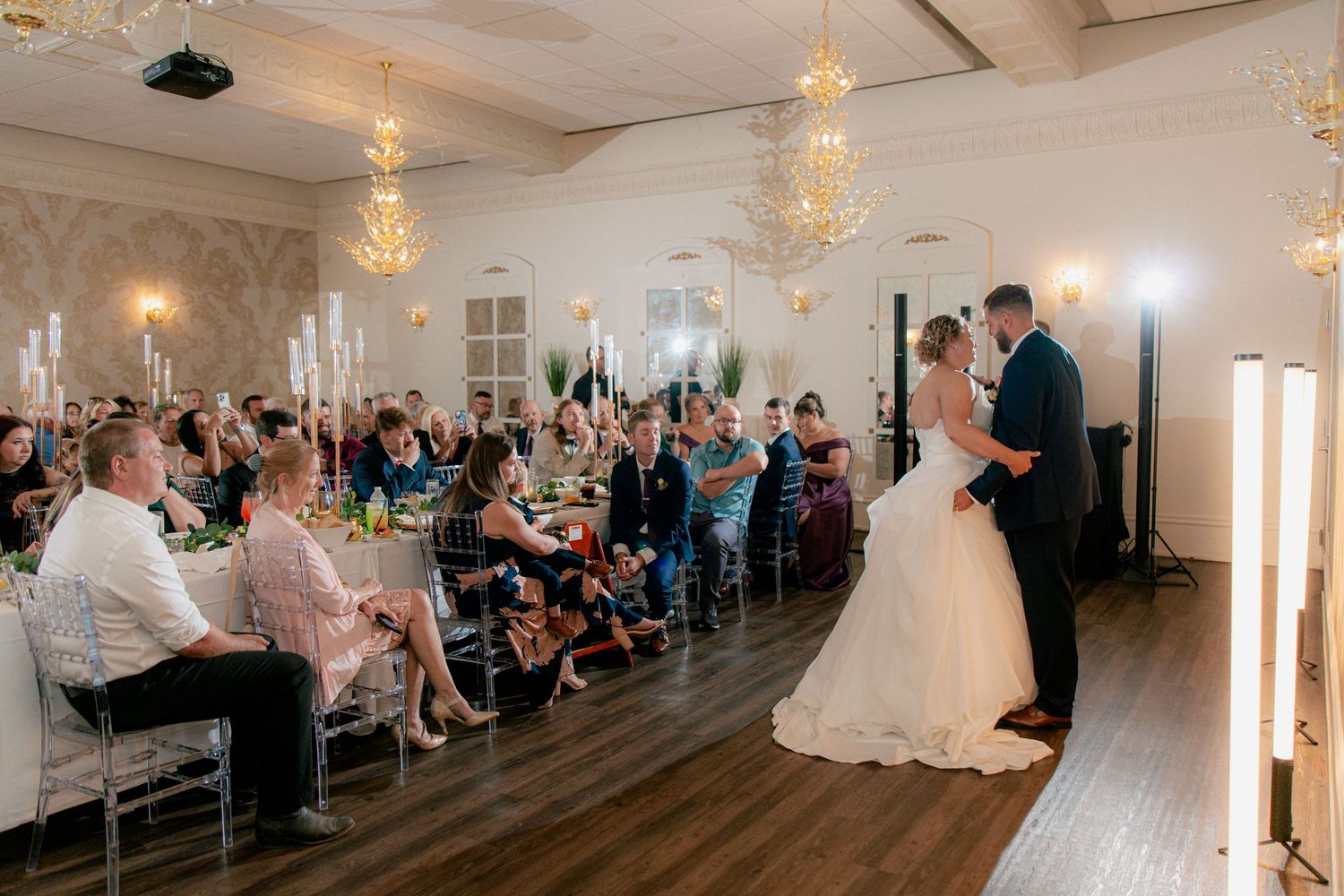 Bride and groom share a romantic first dance before seated wedding reception guests in a chandelier-lit ballroom.