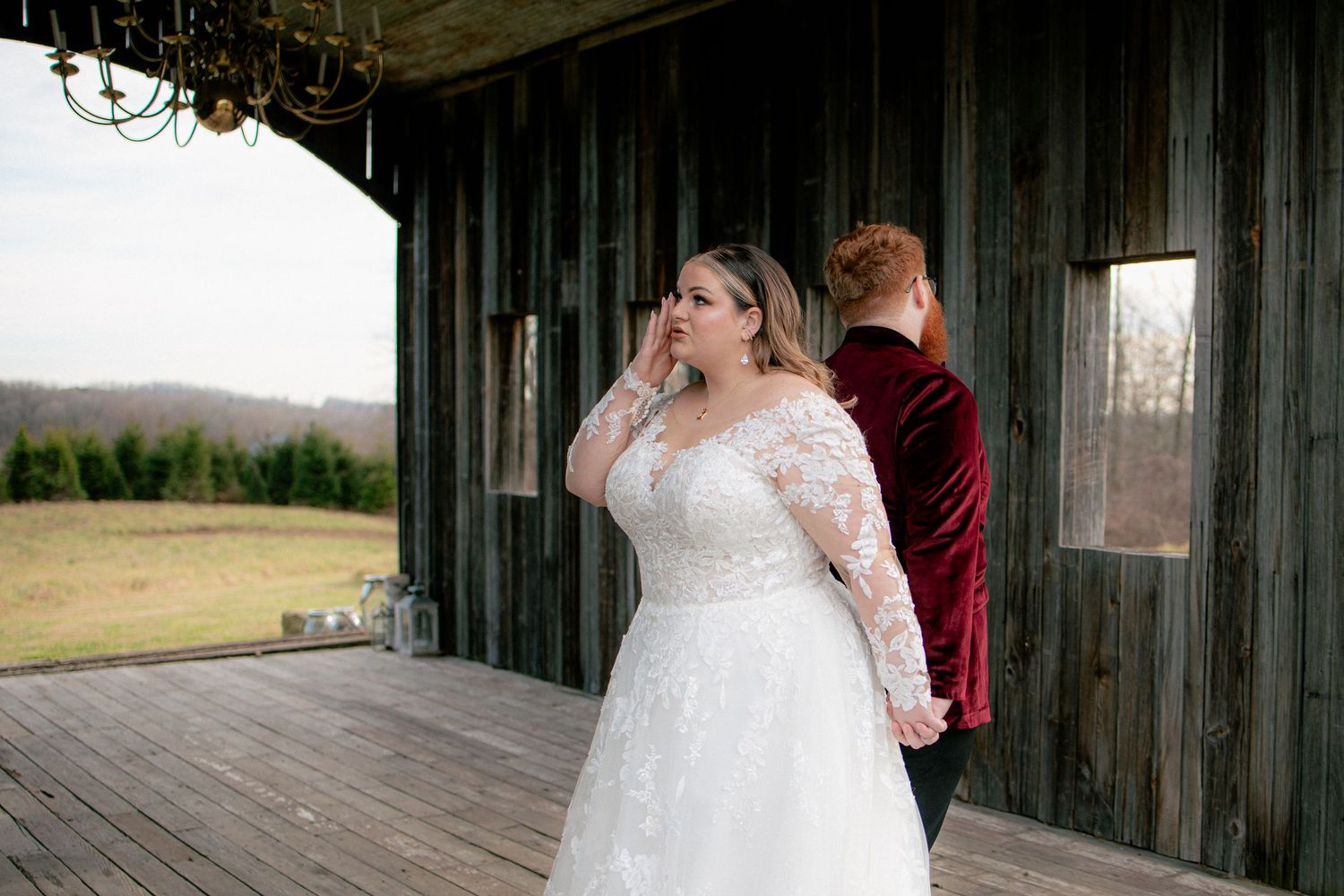 Emotional bride wipes tears during first look moment with groom on rustic barn porch in winter.