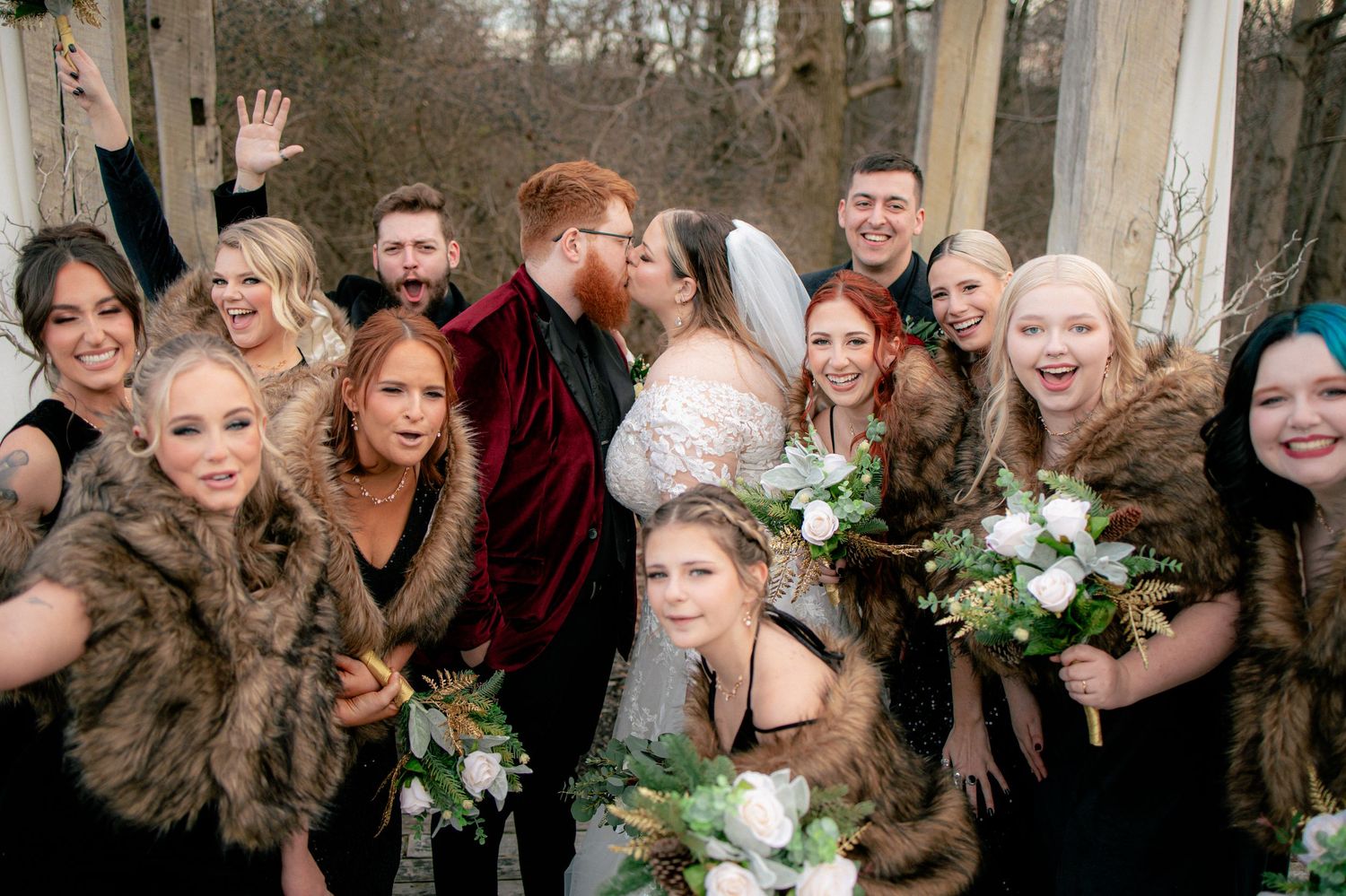 Bride and groom kissing surrounded by wedding party in fur wraps during a winter outdoor celebration.