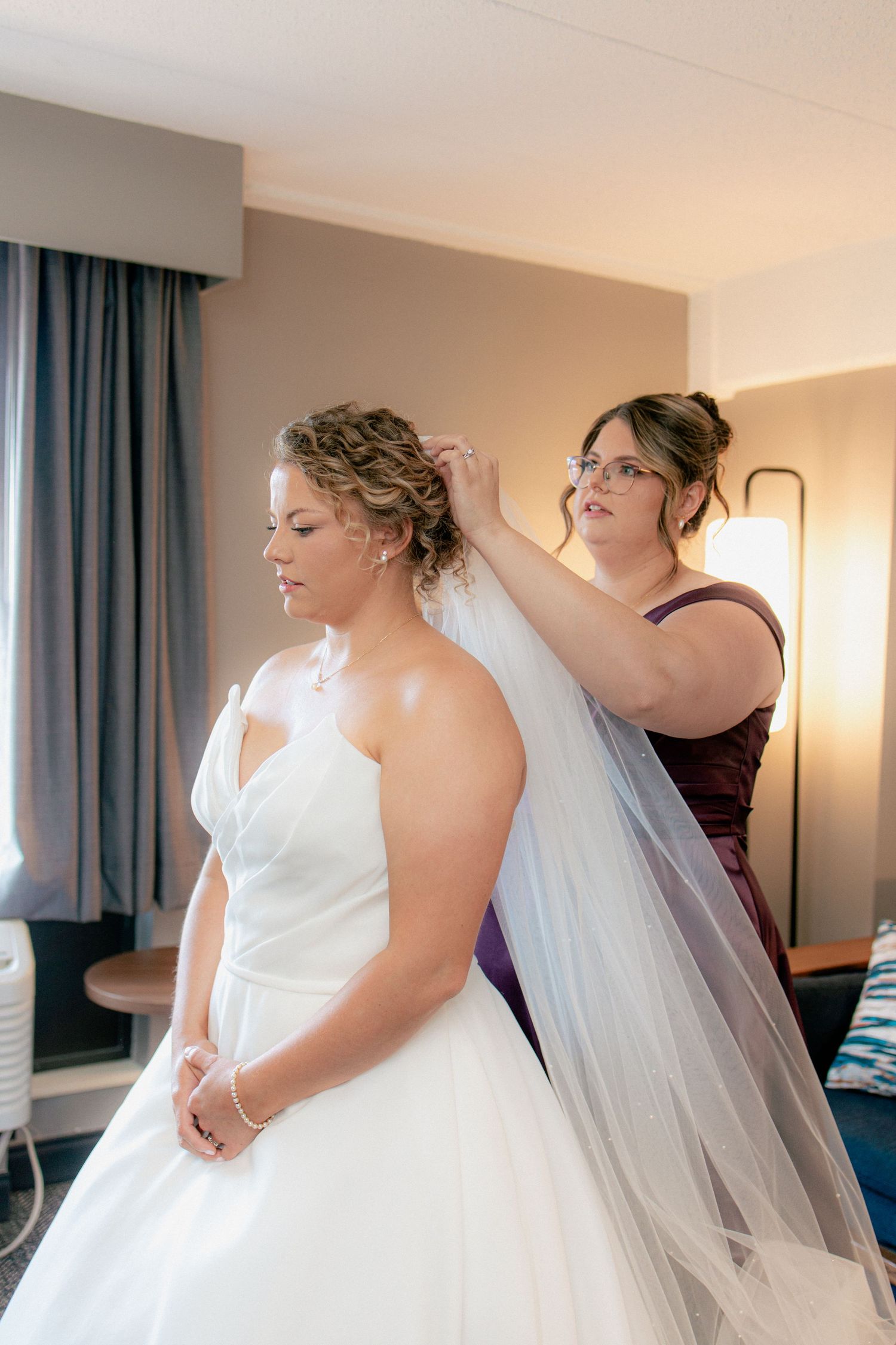 Bridesmaid carefully attaches a long veil to the brides updo hairstyle in a hotel room.