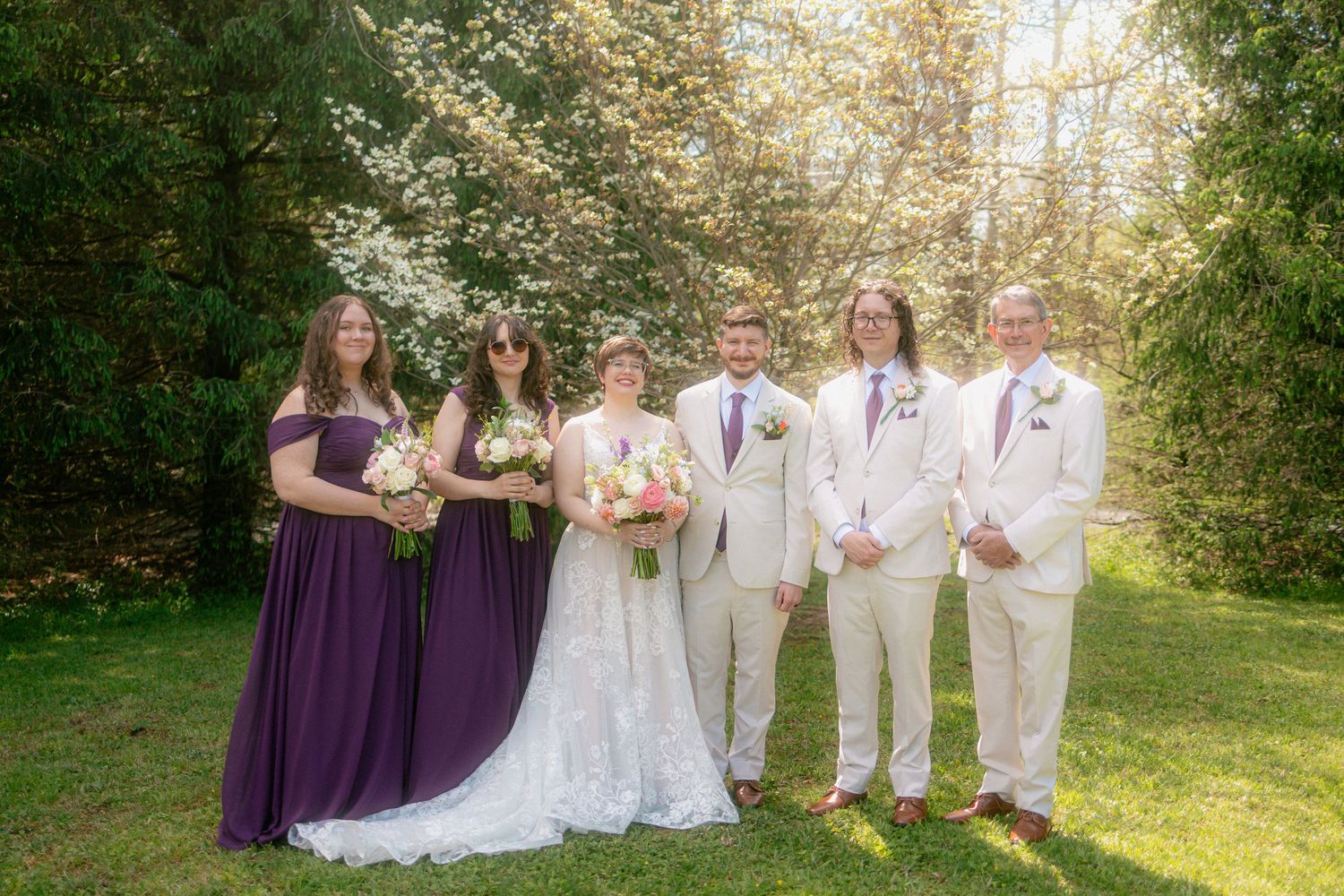 Wedding party posing outdoors in lush green garden, bridesmaids in purple gowns, groomsmen in tan suits, bride in lace dress.