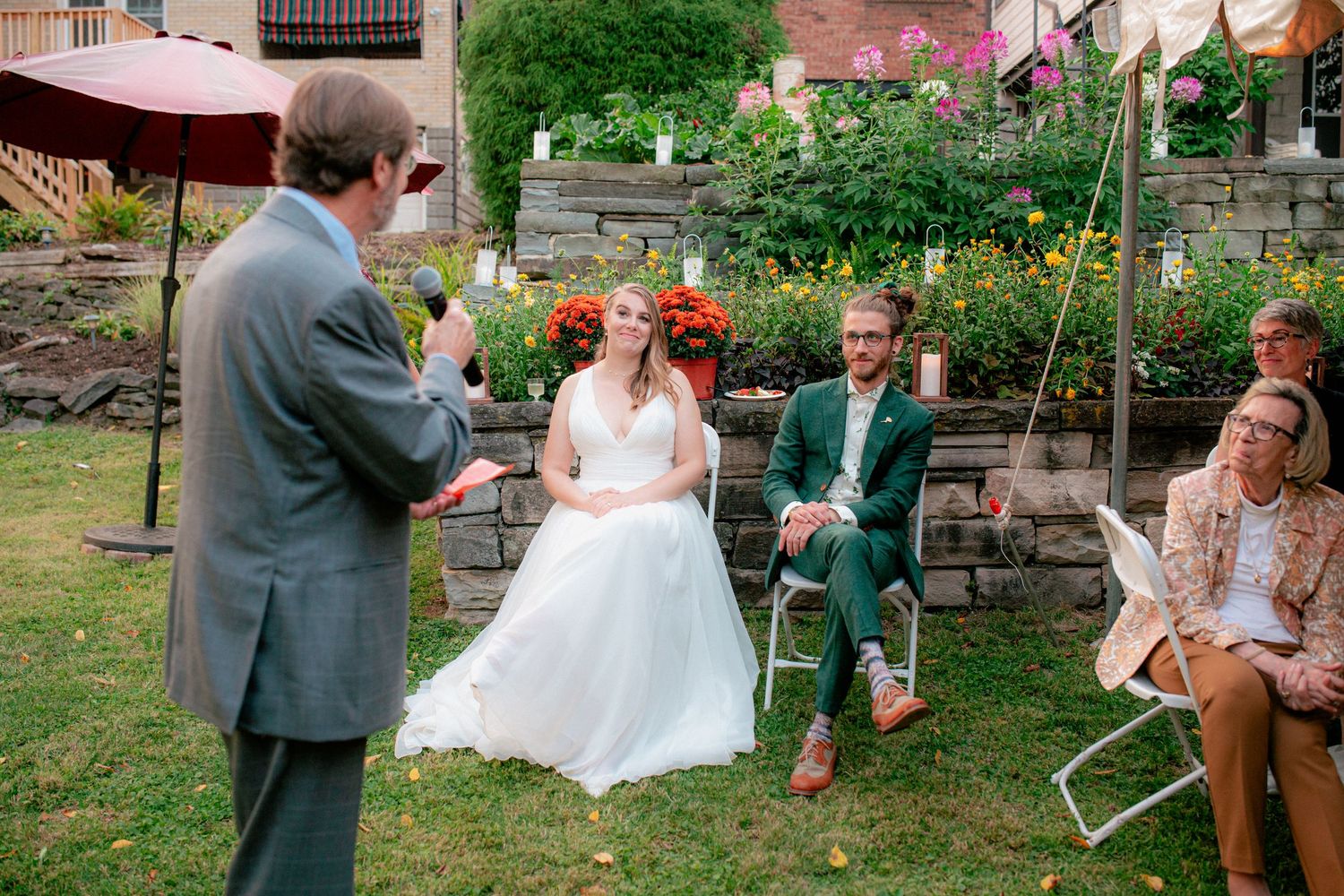 Bride and groom in green suit listen to speech during intimate outdoor garden wedding ceremony.