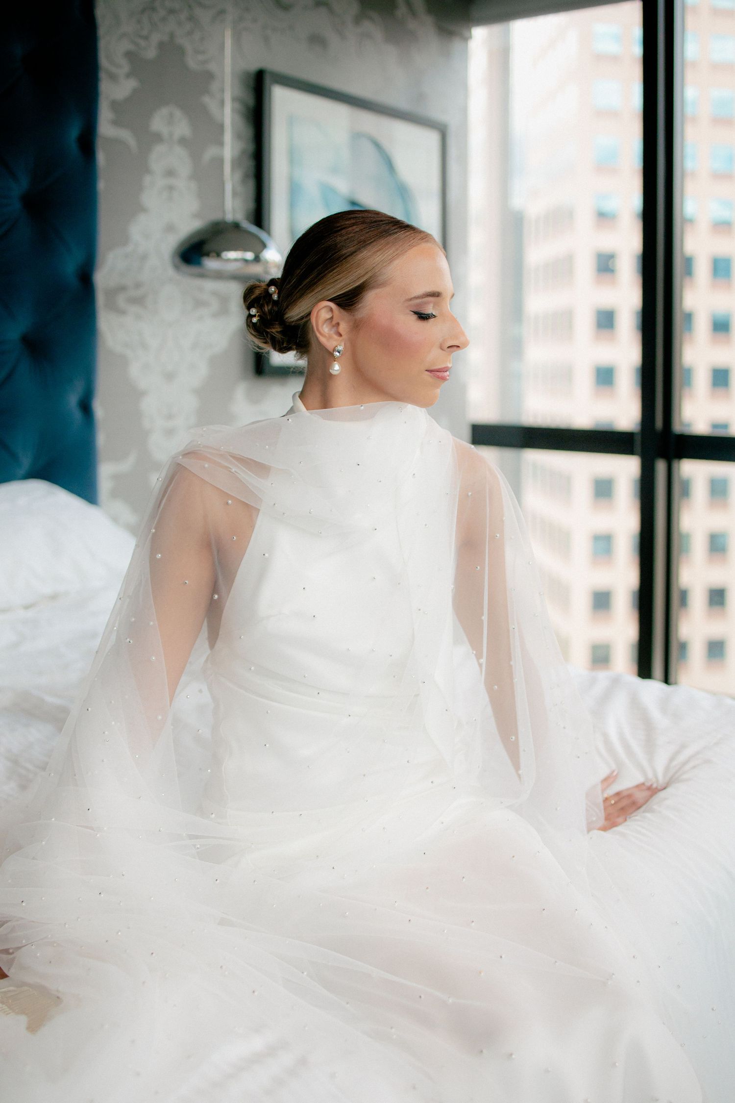 Bride in white halter gown with tulle cape sits on bed near city window, eyes closed, pearl earrings gleaming.