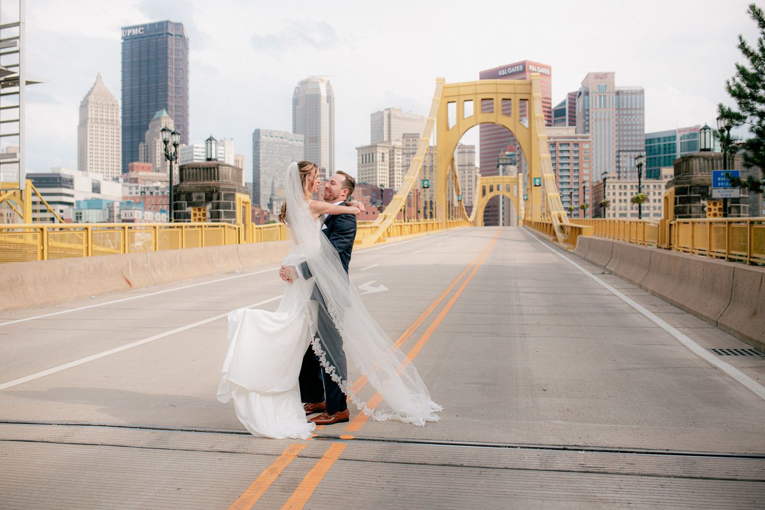 Groom lifts bride in the middle of a Pittsburgh bridge with city skyline in background.