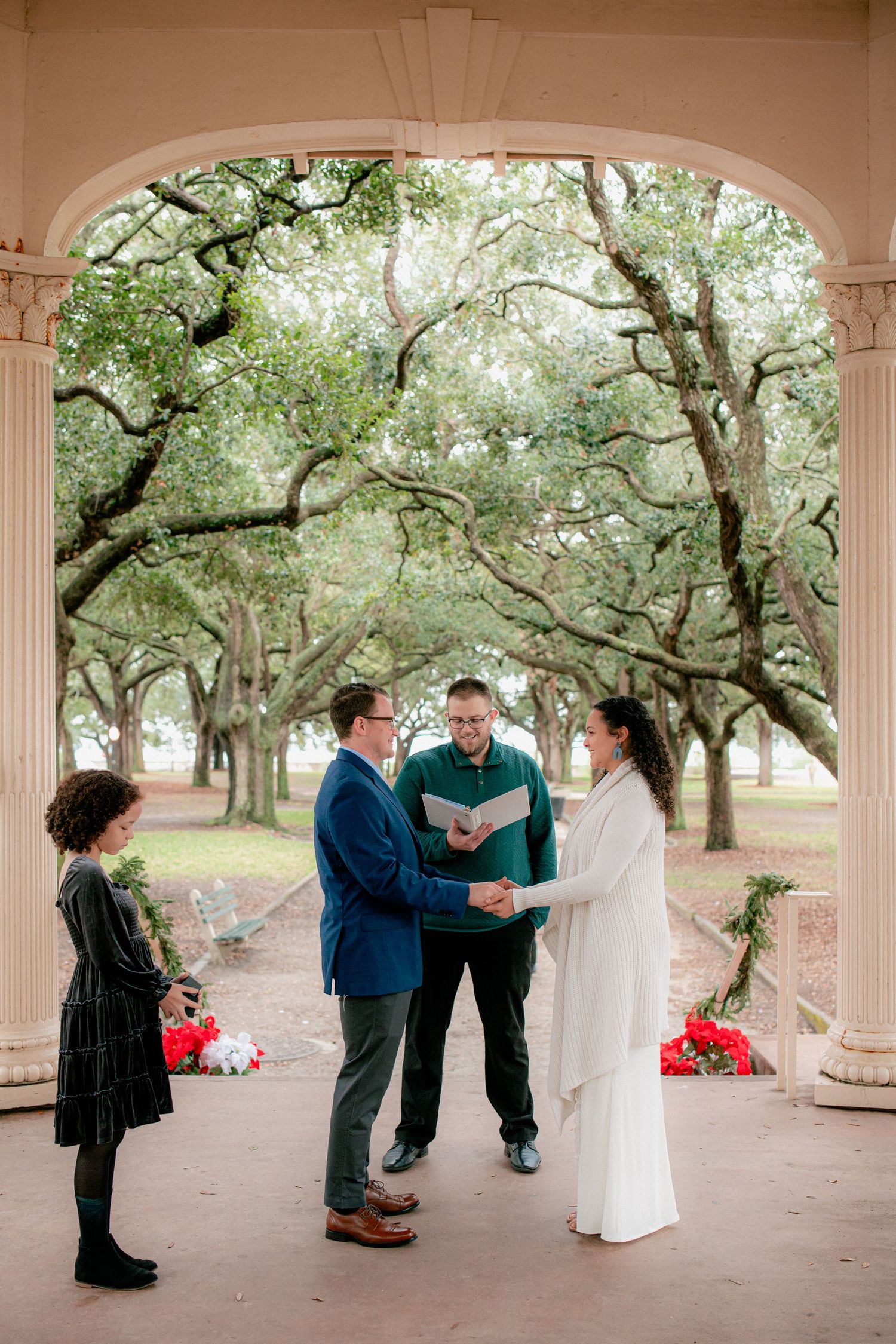 Couple exchanges vows holding hands under grand oak trees at elegant columned outdoor wedding ceremony venue.