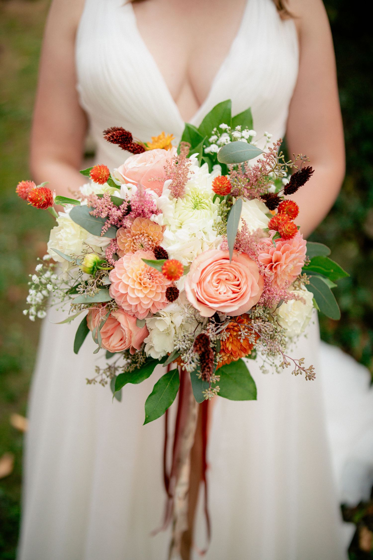 Bride holds lush bridal bouquet with peach roses, dahlias, orange blooms, and greenery with ribbon streamers.