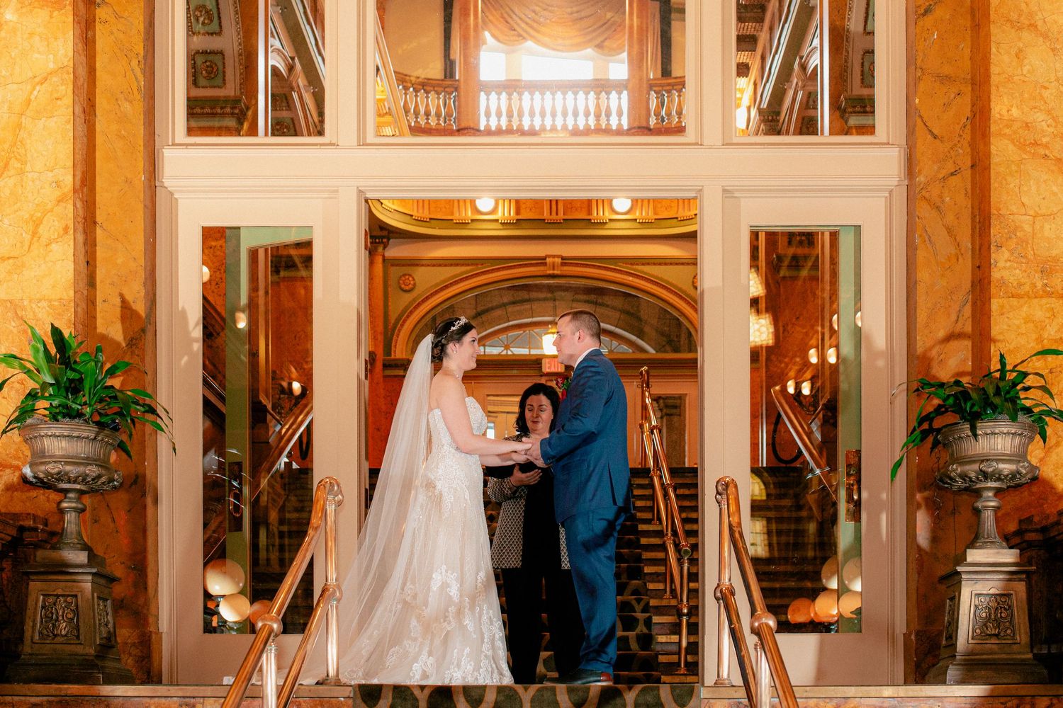 Bride and groom exchange vows at an elegant grand staircase wedding ceremony in a ornate golden venue.