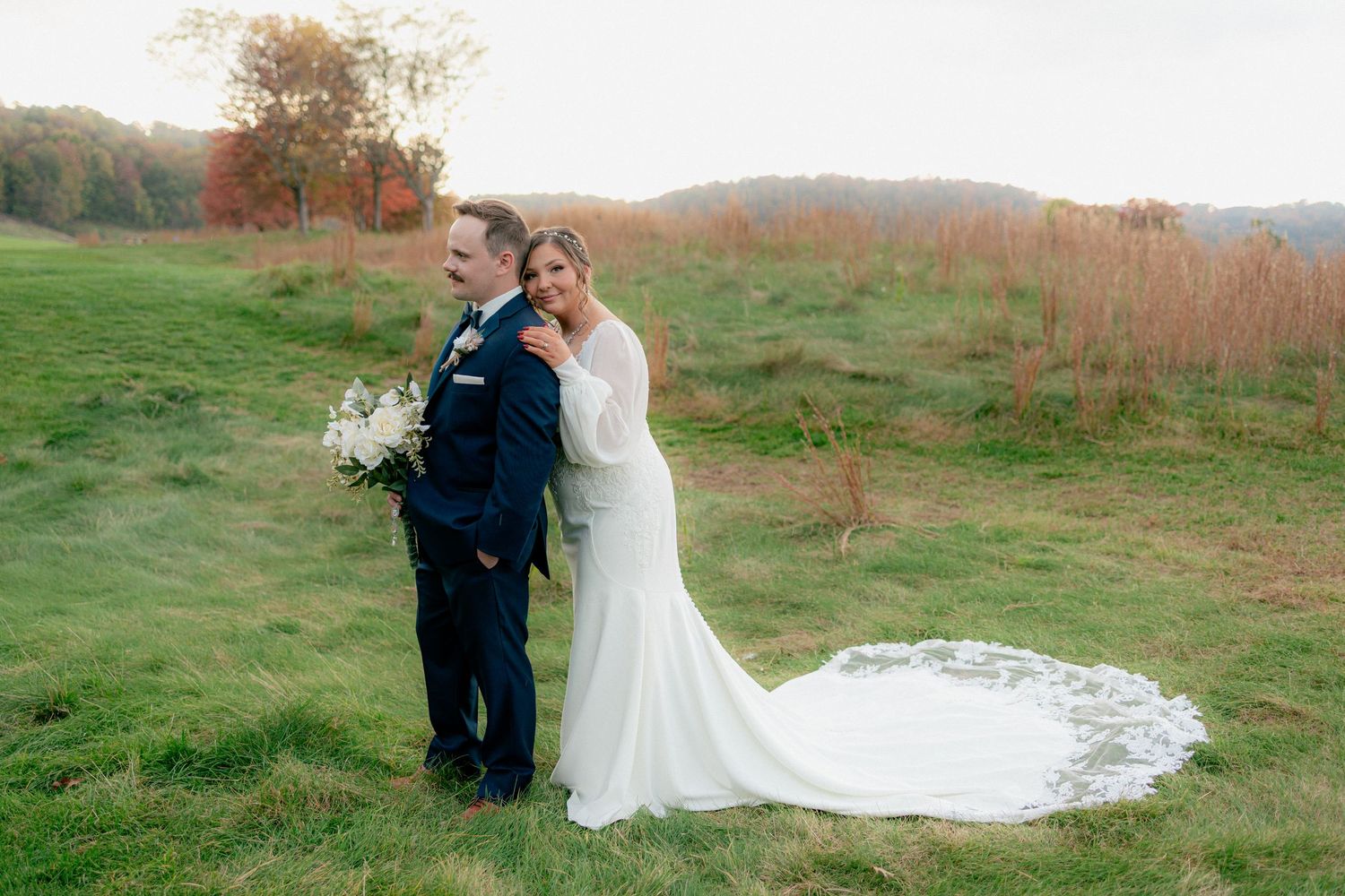 Bride in elegant fitted gown embraces groom from behind in a scenic autumn hillside wedding portrait.
