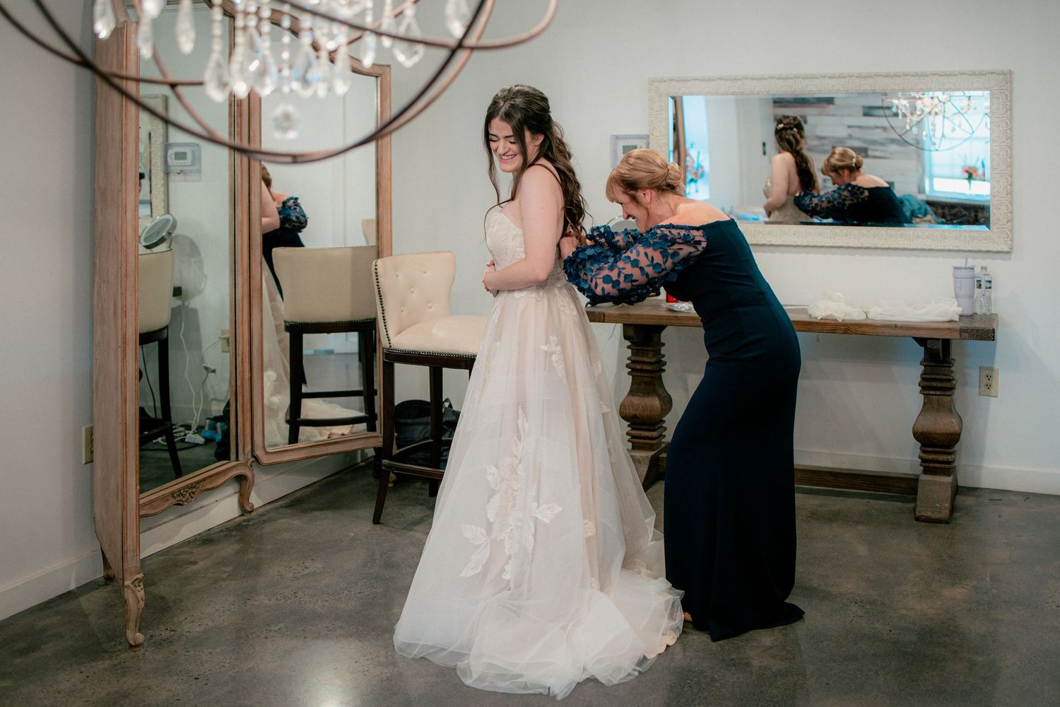 Helper fastening the back of a brides blush wedding gown in a bright dressing room with mirrors.