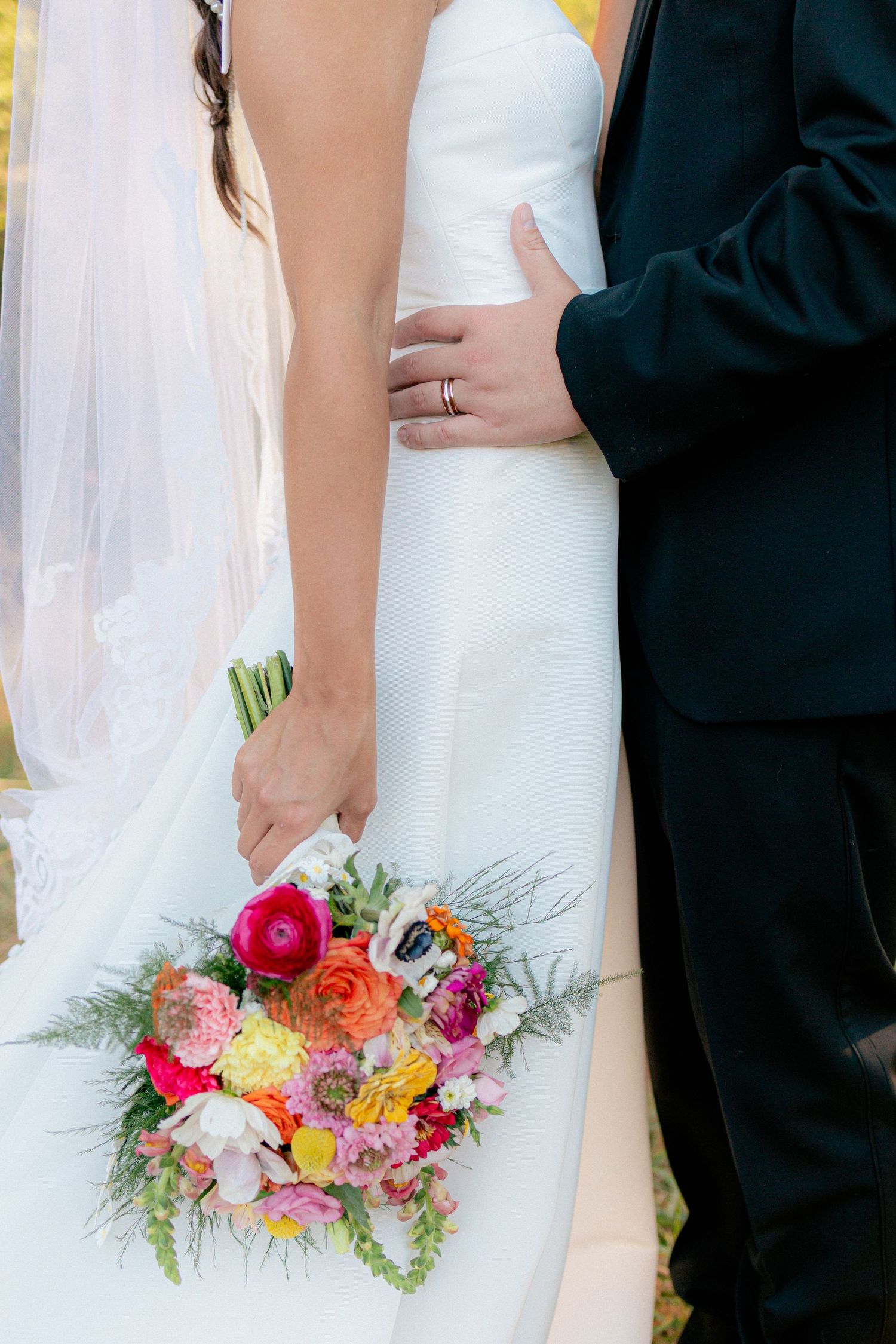 Detail shot of bride and groom standing close, bride holding vibrant multicolored bouquet with rings visible.