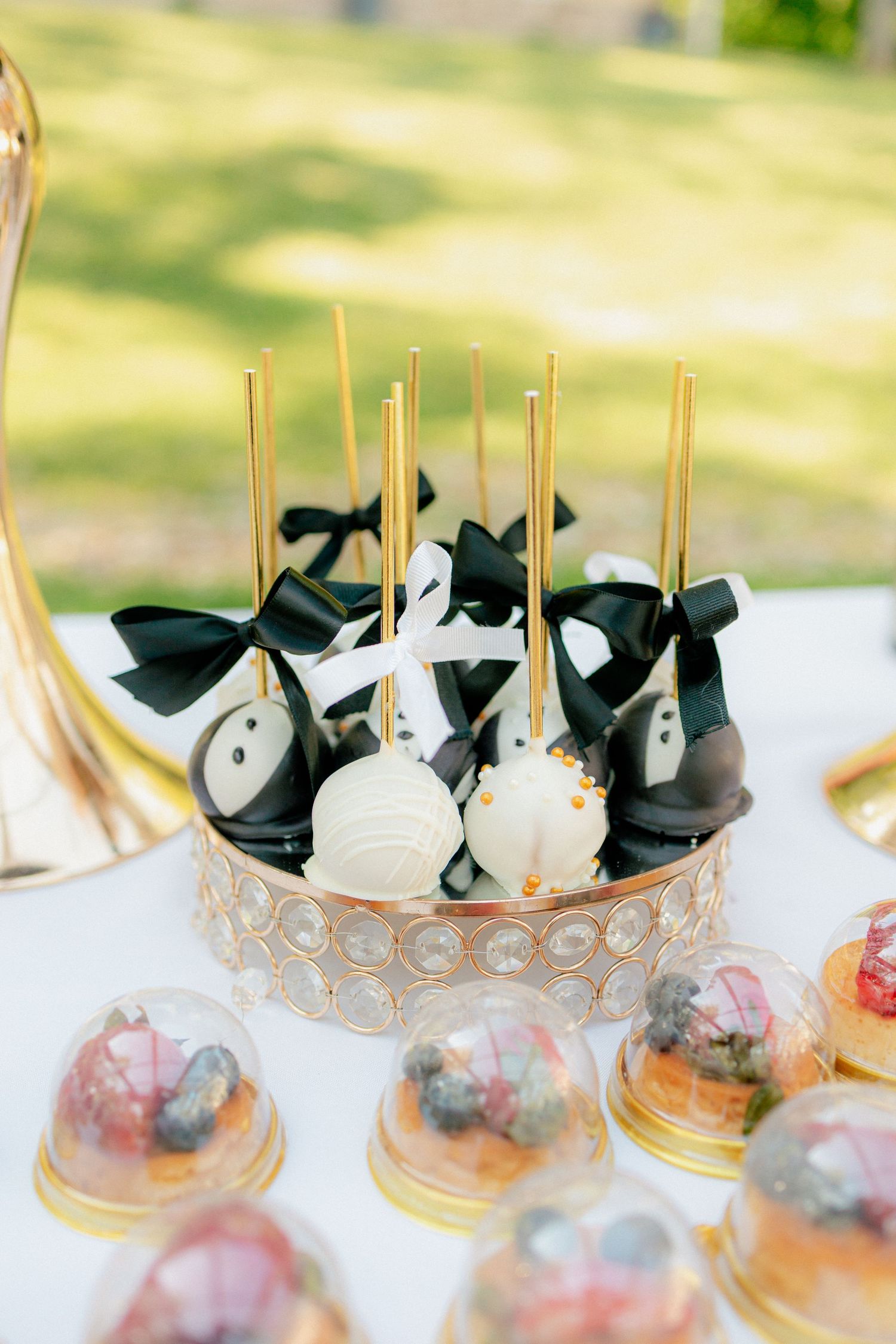 Elegant wedding cake pops decorated with black bow ties and white dresses on a gold stand at an outdoor reception.
