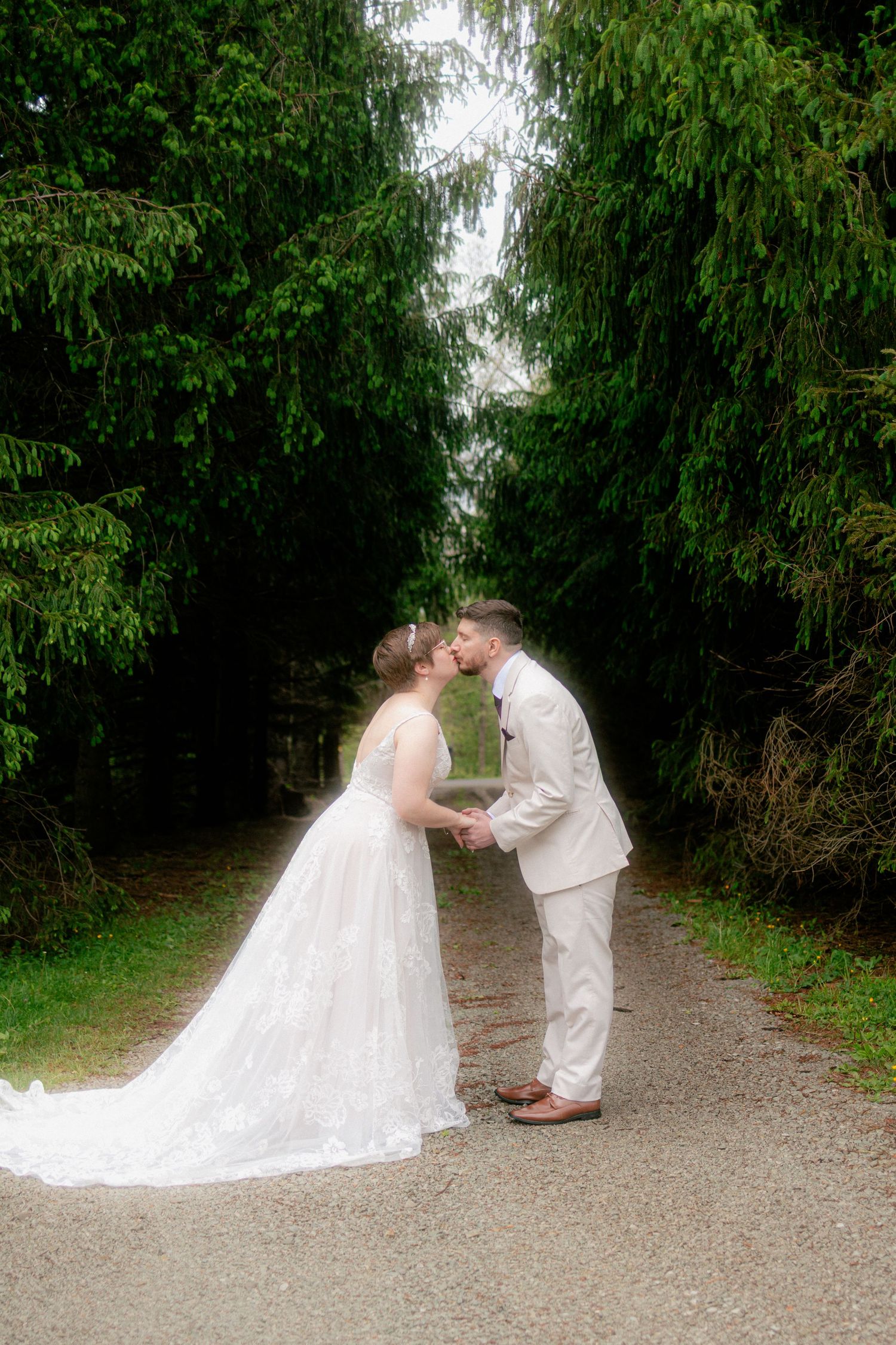 Bride and groom share a kiss on a tree-lined path, bride in white gown with train, groom in beige suit.
