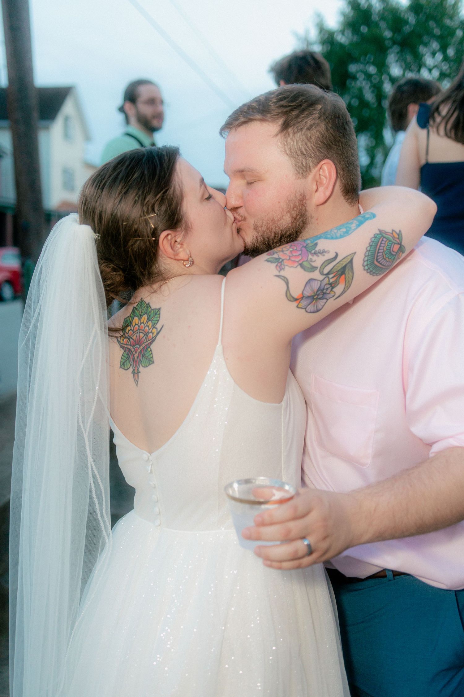 Tattooed bride and groom share a kiss in wedding attire, bride wearing white gown and veil outdoors.