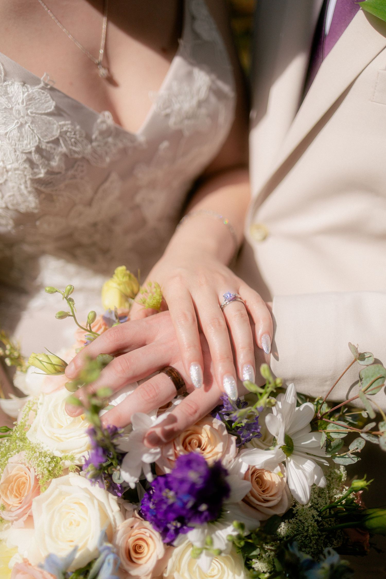 Bride and groom hands resting on wedding bouquet with purple and white flowers, showcasing elegant engagement ring.