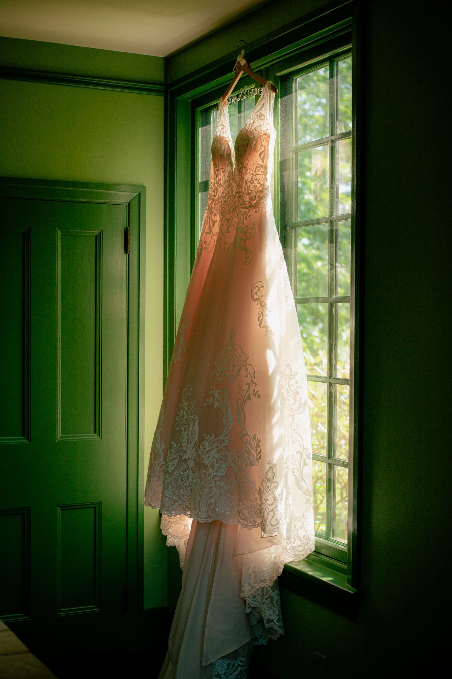 Elegant lace wedding dress hanging in a window with green walls and natural light filtering through.
