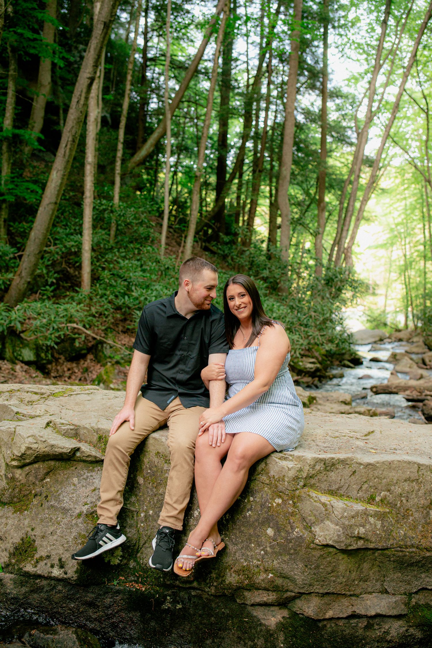Couple sitting on rocks by a forest stream, smiling at each other during engagement session.