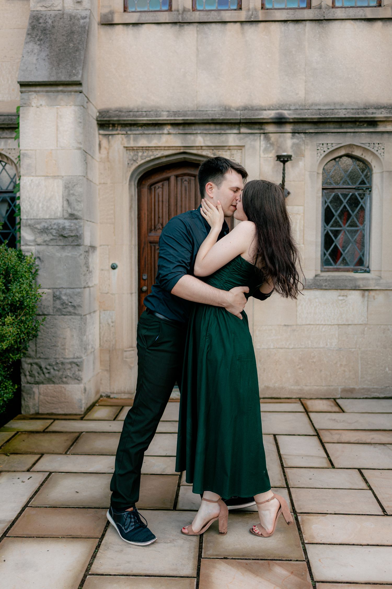 Couple sharing a passionate kiss outside a historic stone building, woman in green dress and man in dark outfit.