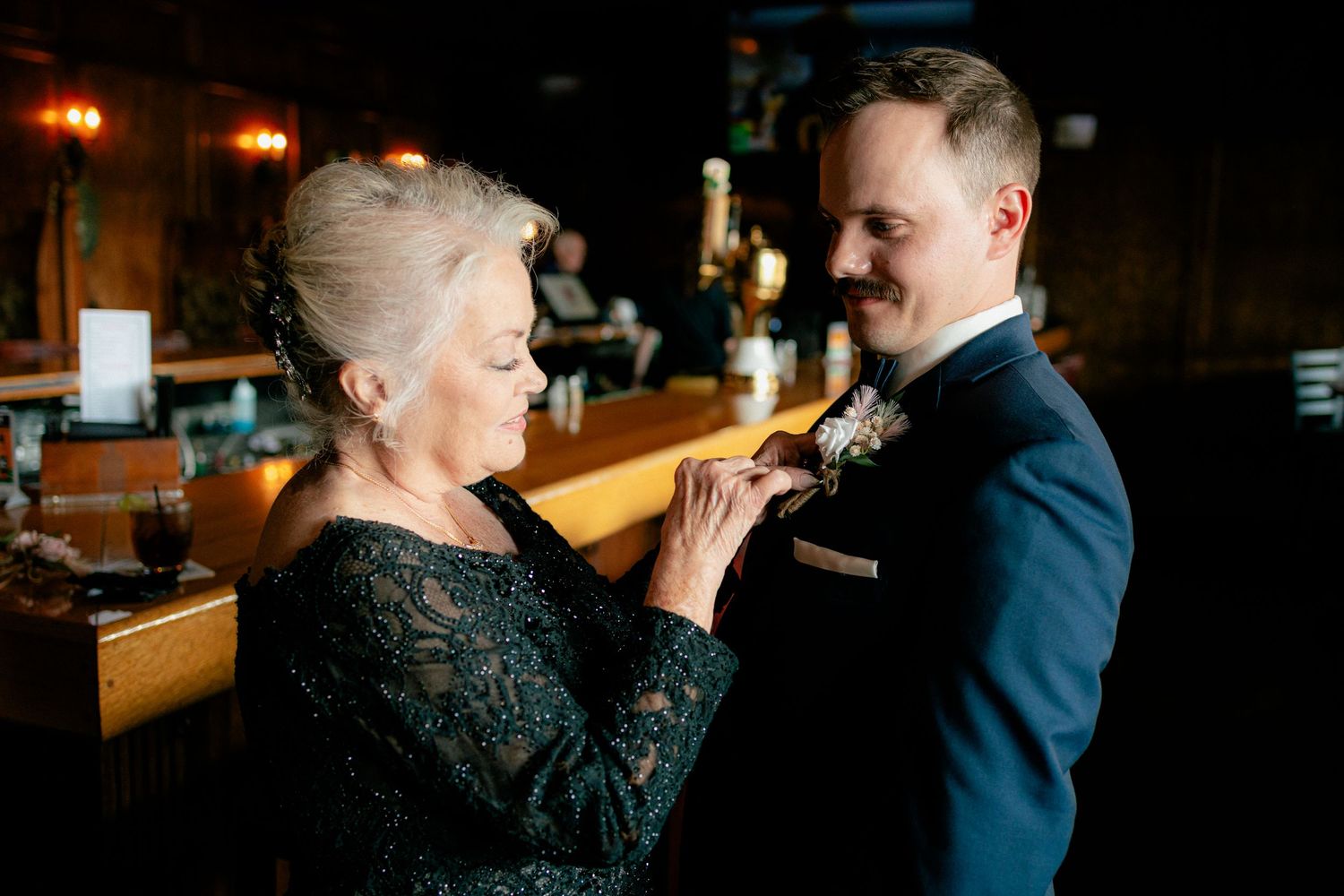 Elderly woman pinning a boutonniere on a groom in a navy suit at a wedding venue bar.
