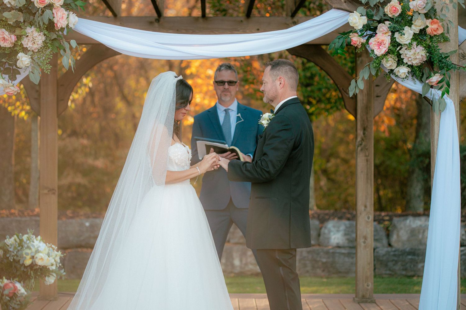 Bride and groom hold hands at an outdoor altar decorated with pink flowers and draped fabric.