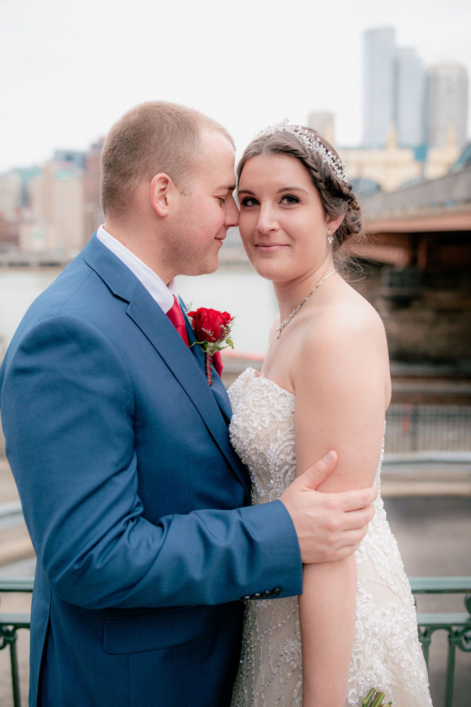 Bride in beaded gown and groom in blue suit embrace closely with Pittsburgh bridge backdrop.