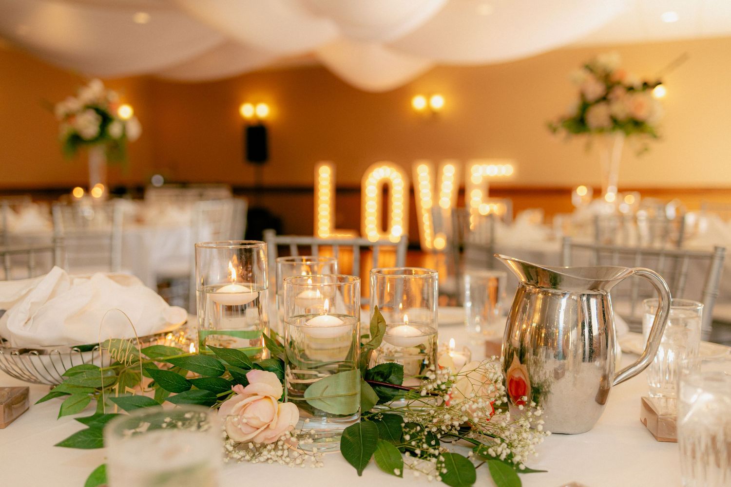 Wedding reception table centerpiece with candles, greenery, and illuminated LOVE marquee letters in background.
