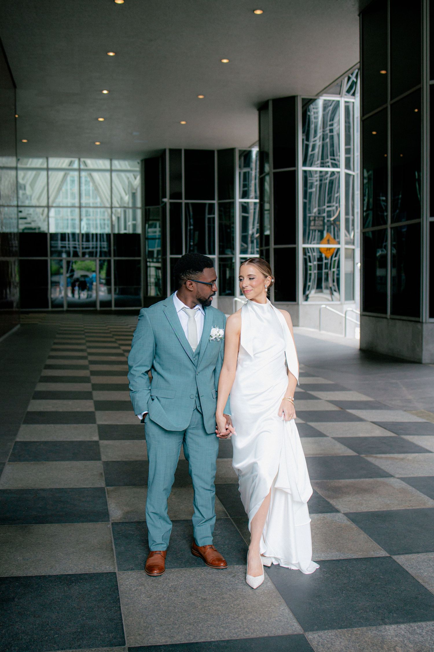 Interracial couple in wedding attire hold hands inside modern building with checkered floor, groom in light blue suit.