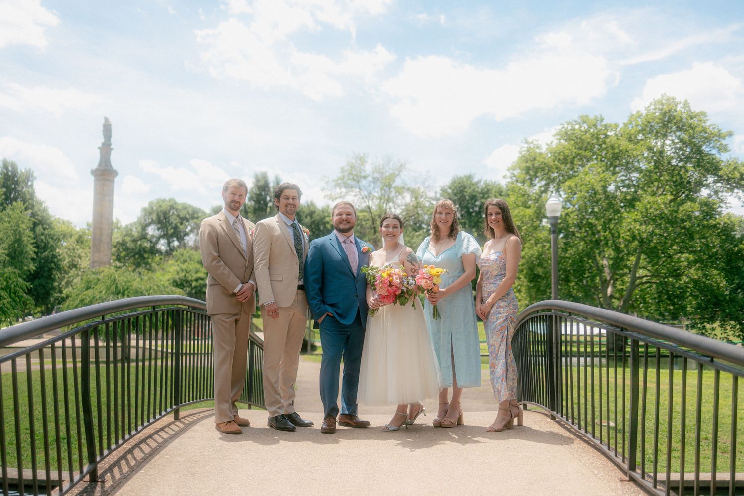 Wedding party of six poses on a bridge in a lush park, bride holding colorful bouquet, sunny day.